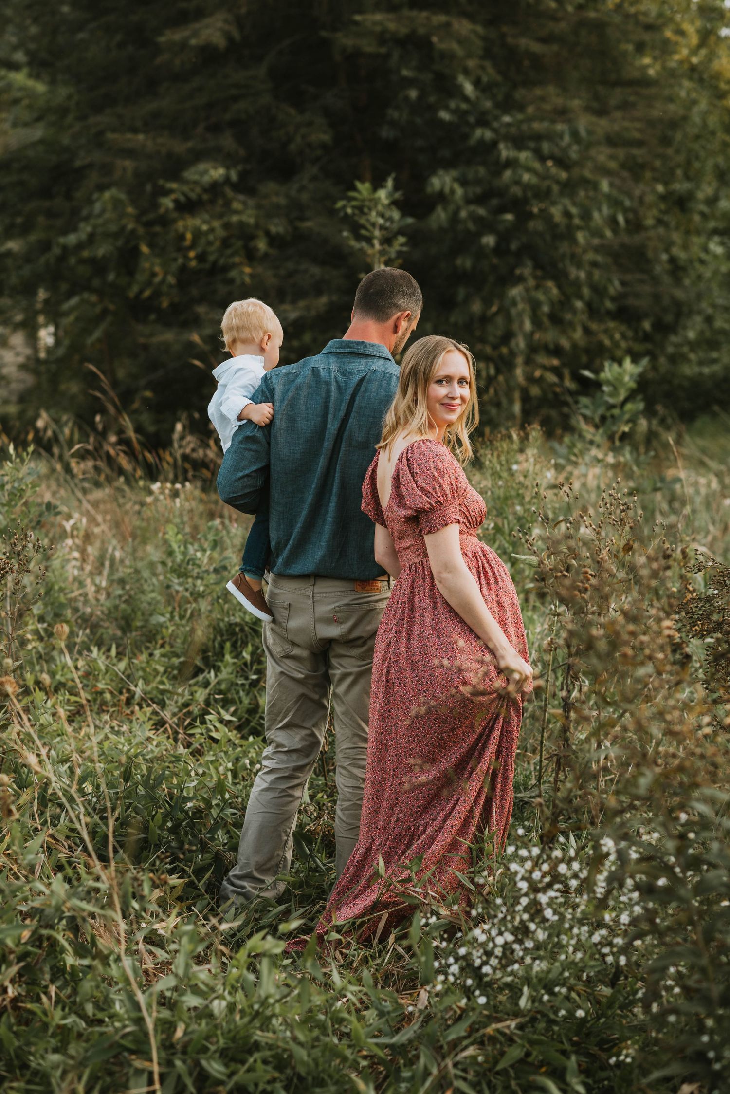 A woman in a red floral dress looks back while her partner carries their toddler in a meadow.