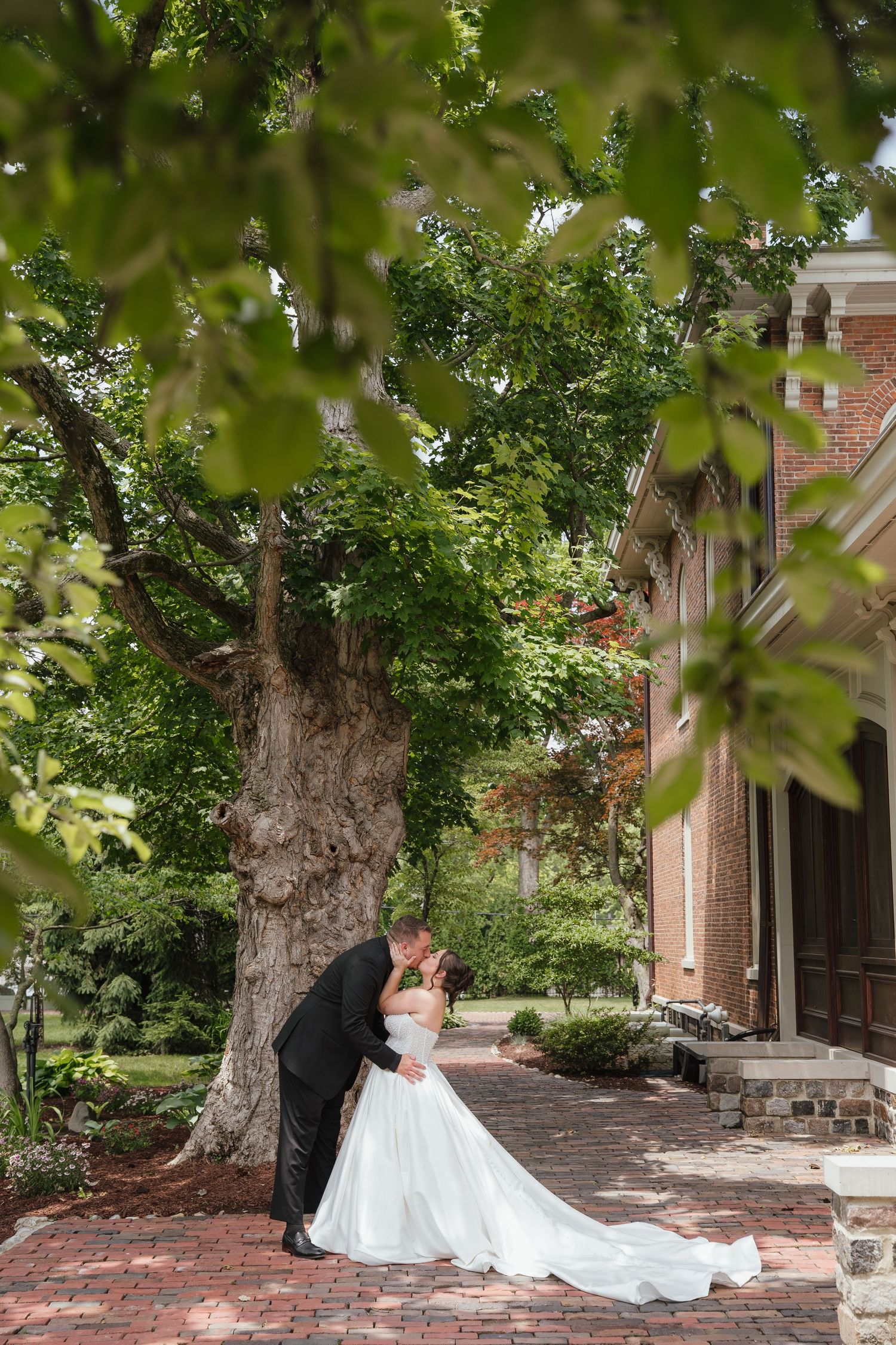 Bride and groom sharing a romantic kiss beneath a large tree outside a historic brick building.
