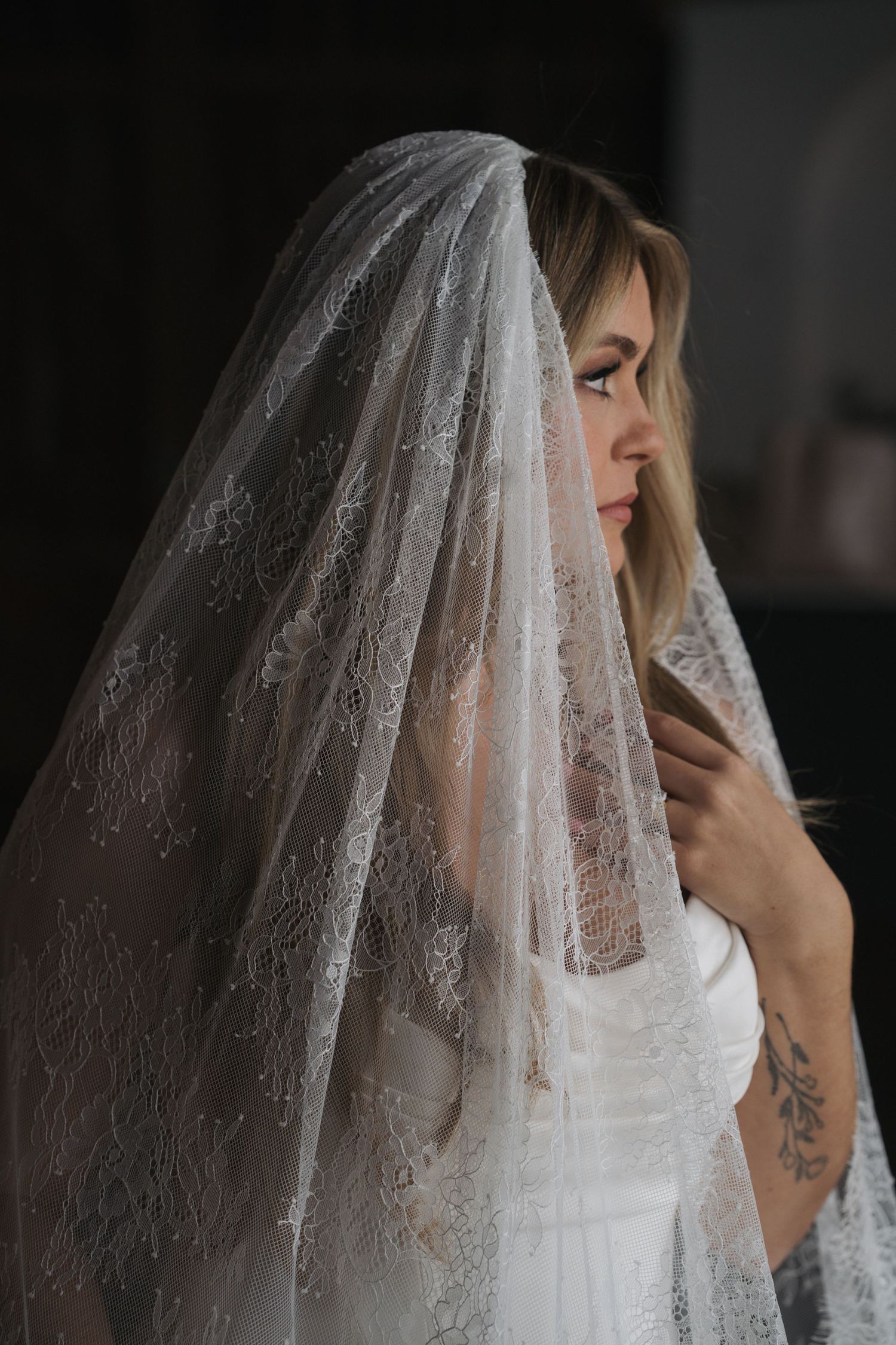 Bride wearing an elegant lace wedding veil with floral details, looking down in soft light.
