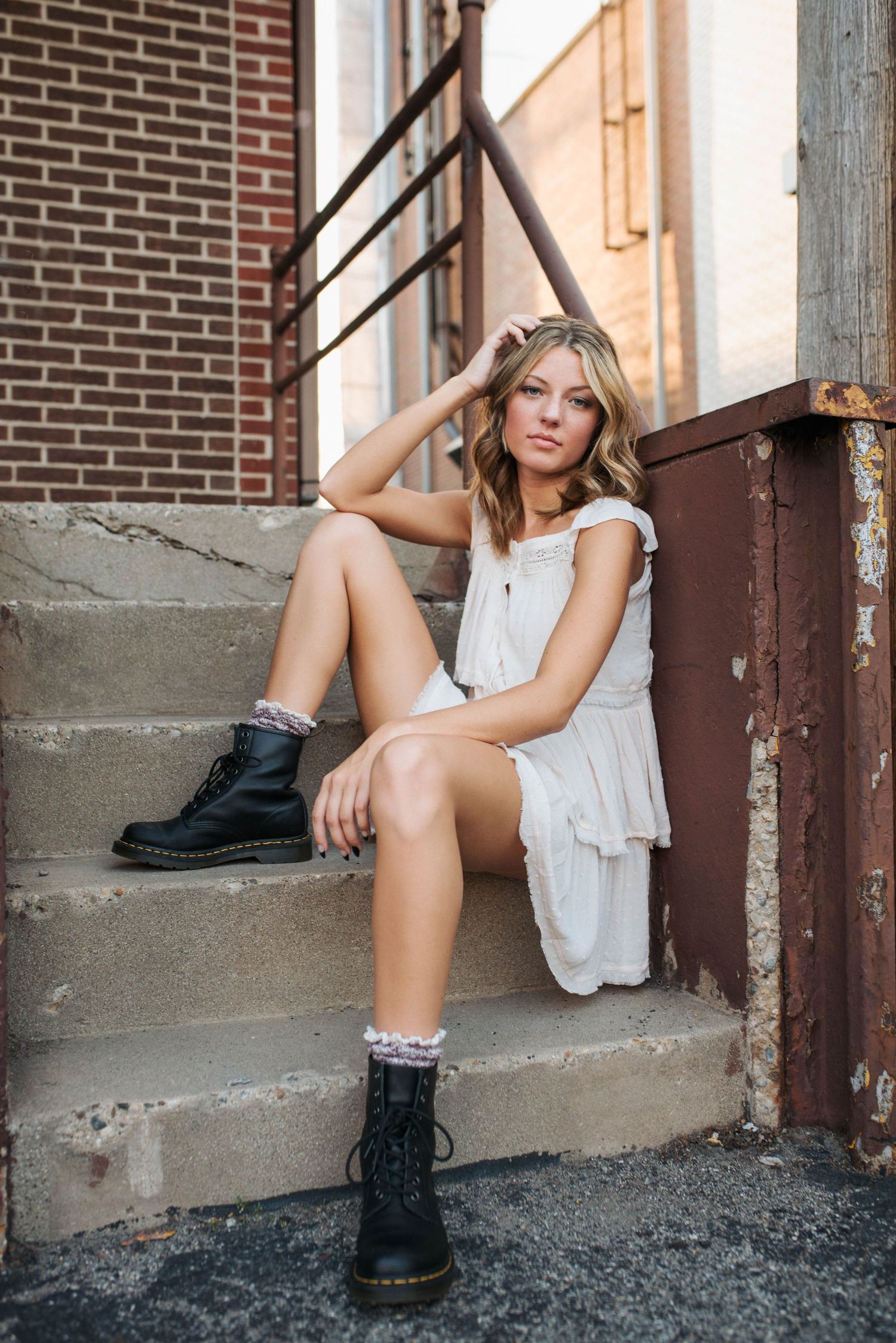 Blonde woman in white dress and black combat boots sitting on outdoor concrete steps.