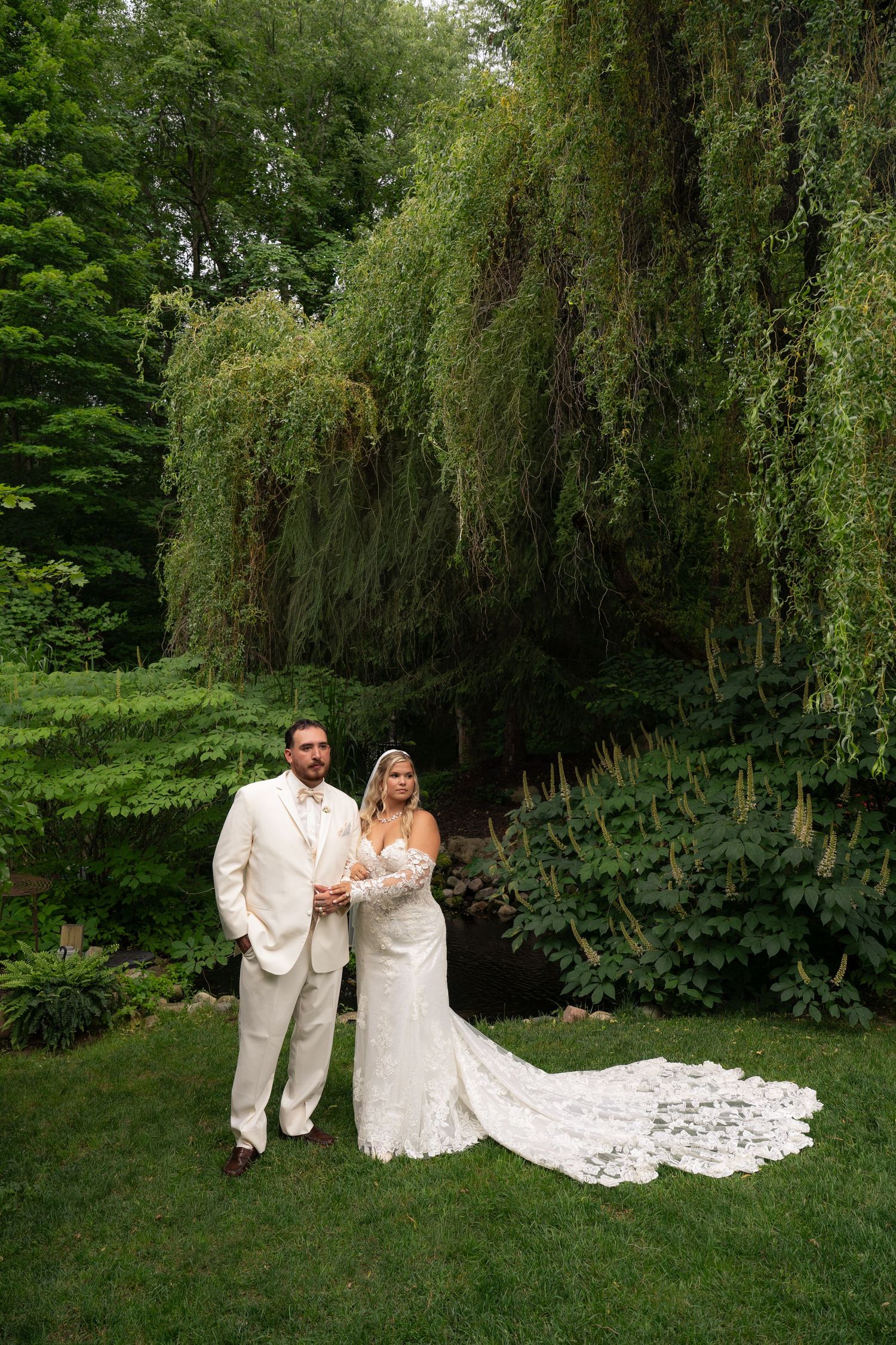 Bride and groom in ivory attire posing together beneath a large weeping willow tree outdoors.