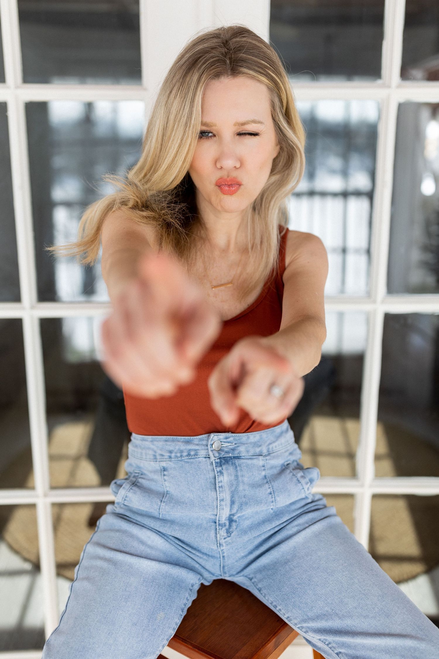 Blonde woman in red tank top and jeans pointing both fingers at camera with pursed lips.