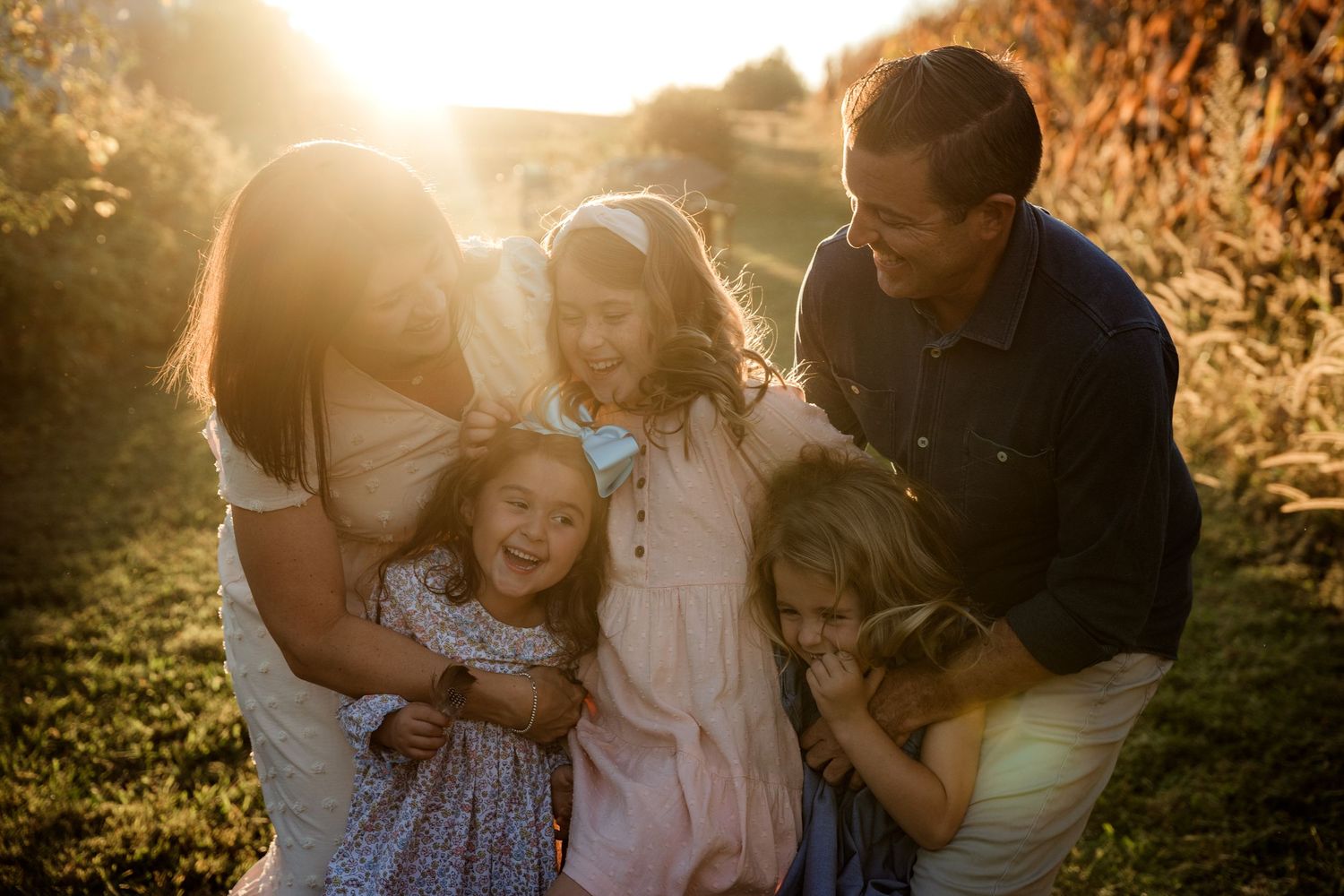 A happy family with three young daughters shares a warm embrace in golden sunlight.