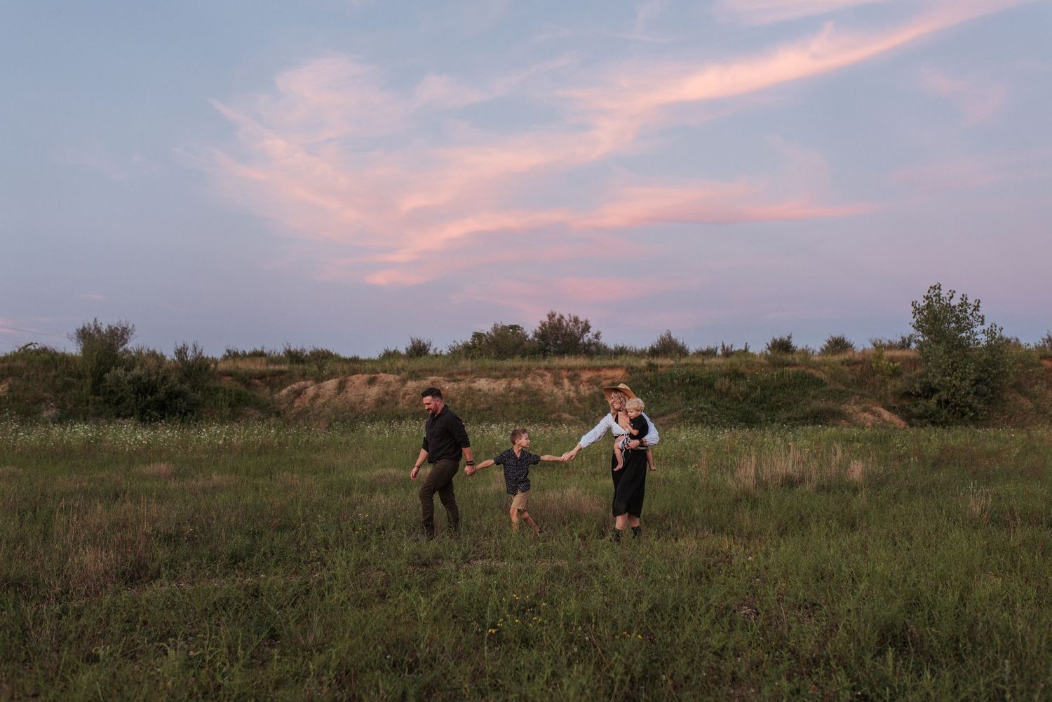 A family of three holding hands while walking through an open green field at sunset.