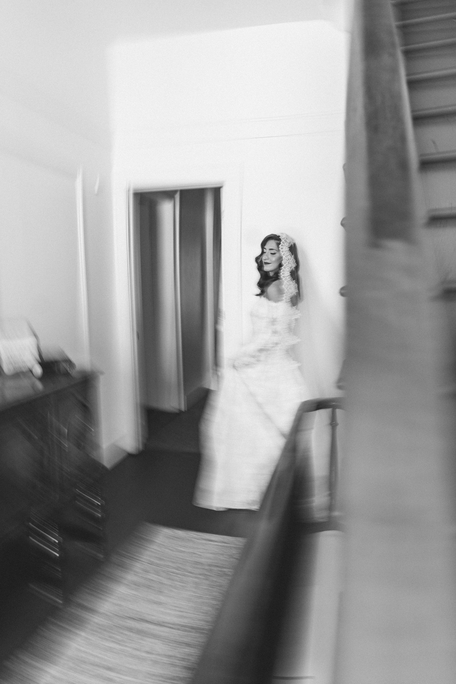 Artistic blurred black and white photo of a bride in a lace gown standing near a staircase.