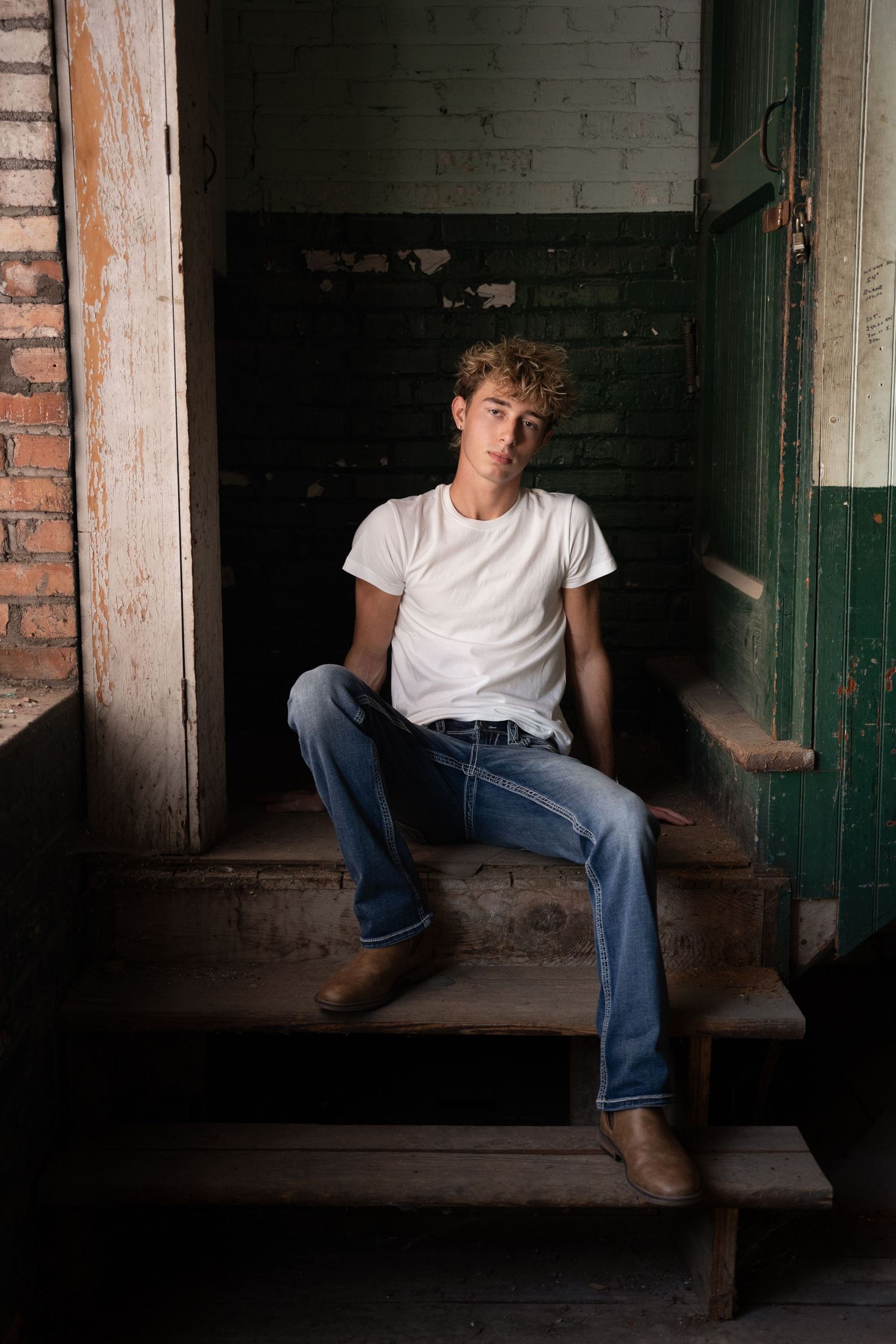 Young man in white t-shirt and jeans sitting on wooden steps in rustic building.