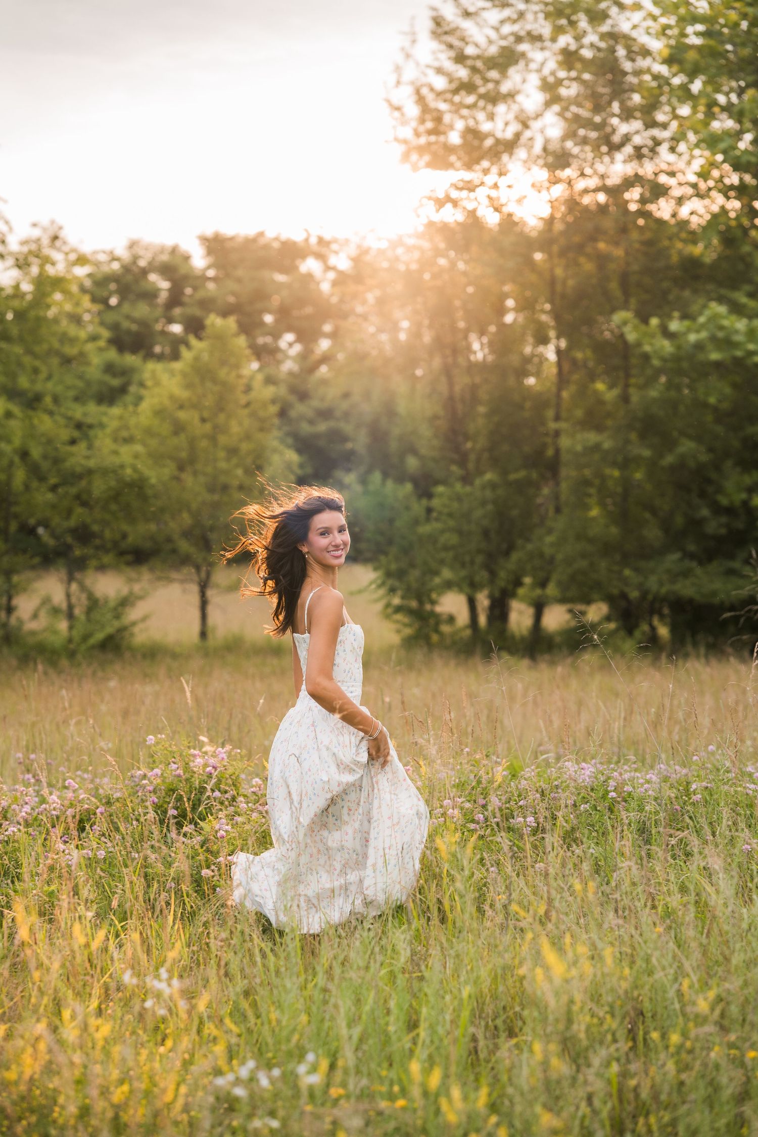 Joyful brunette woman in white lace dress twirling in sunlit meadow with trees in background.