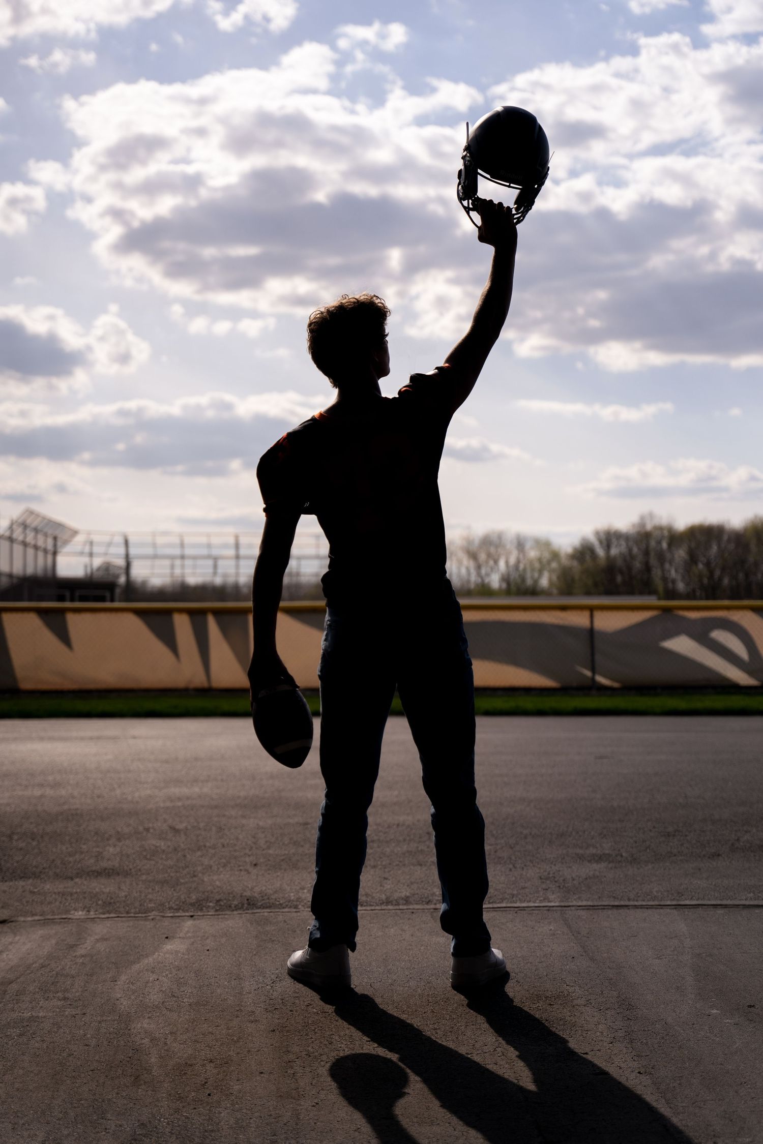 Silhouette of a football player raising helmet triumphantly against a cloudy sky on a sports field.