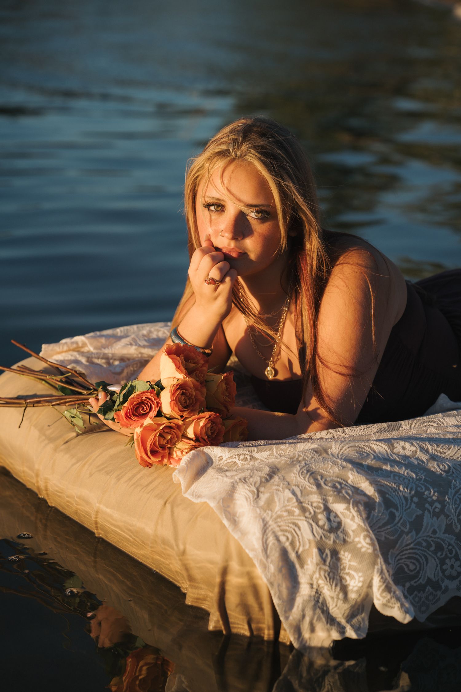 Blonde woman lying on a white lace float in water, holding roses in warm golden sunset light.