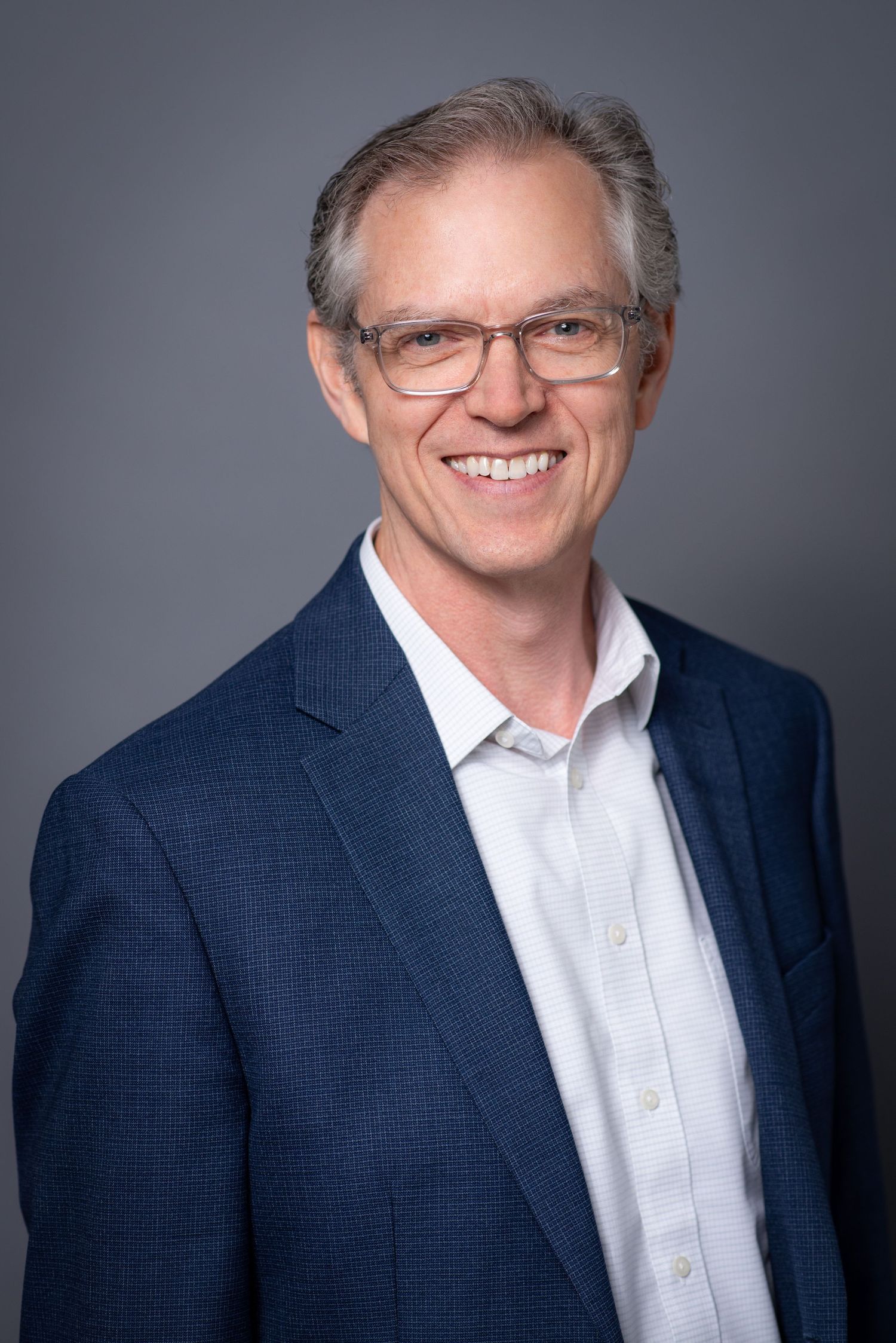 Smiling middle-aged man with glasses wearing a blue blazer and white shirt against a gray background.