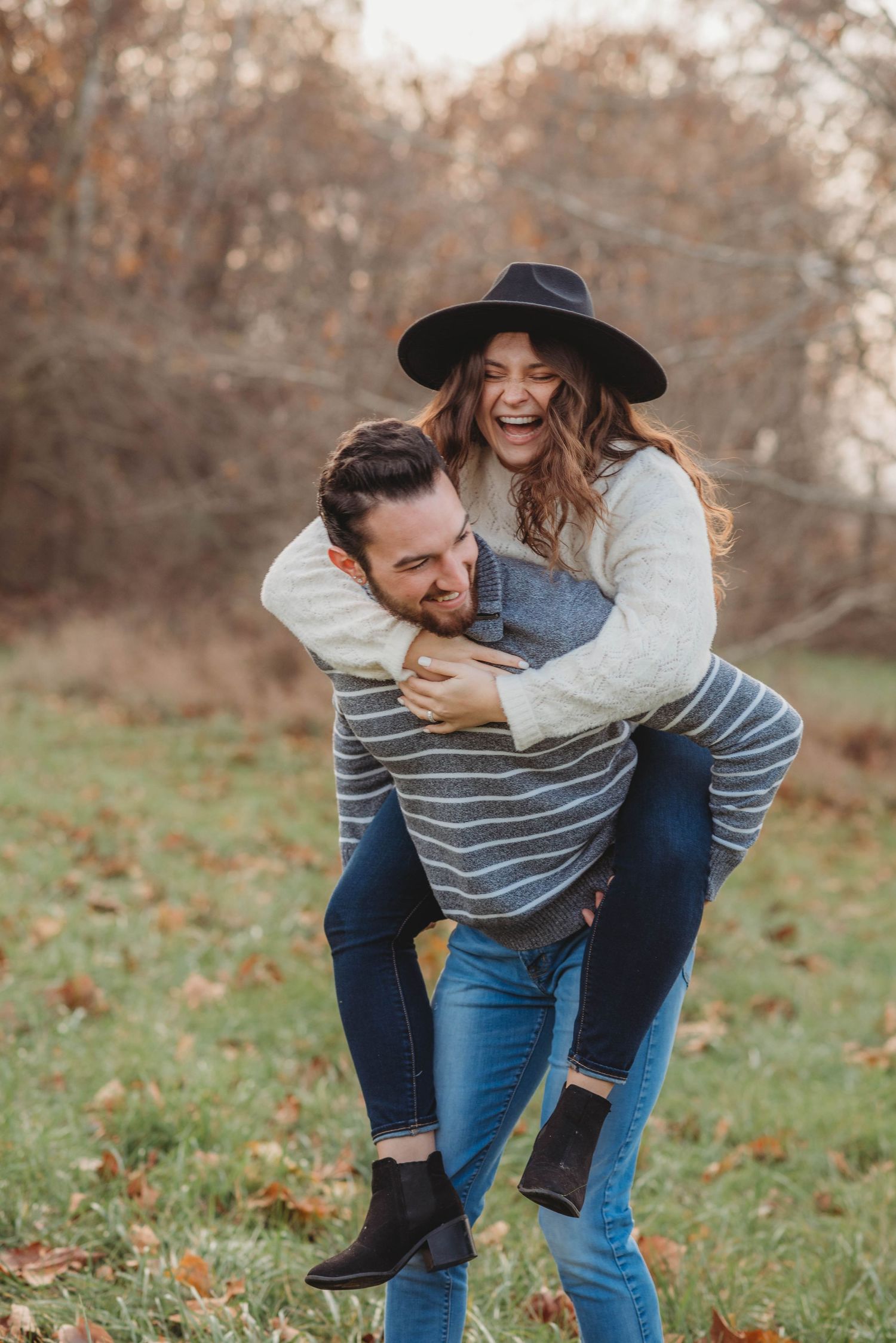 Man giving laughing woman a piggyback ride in an autumn field with fallen leaves.