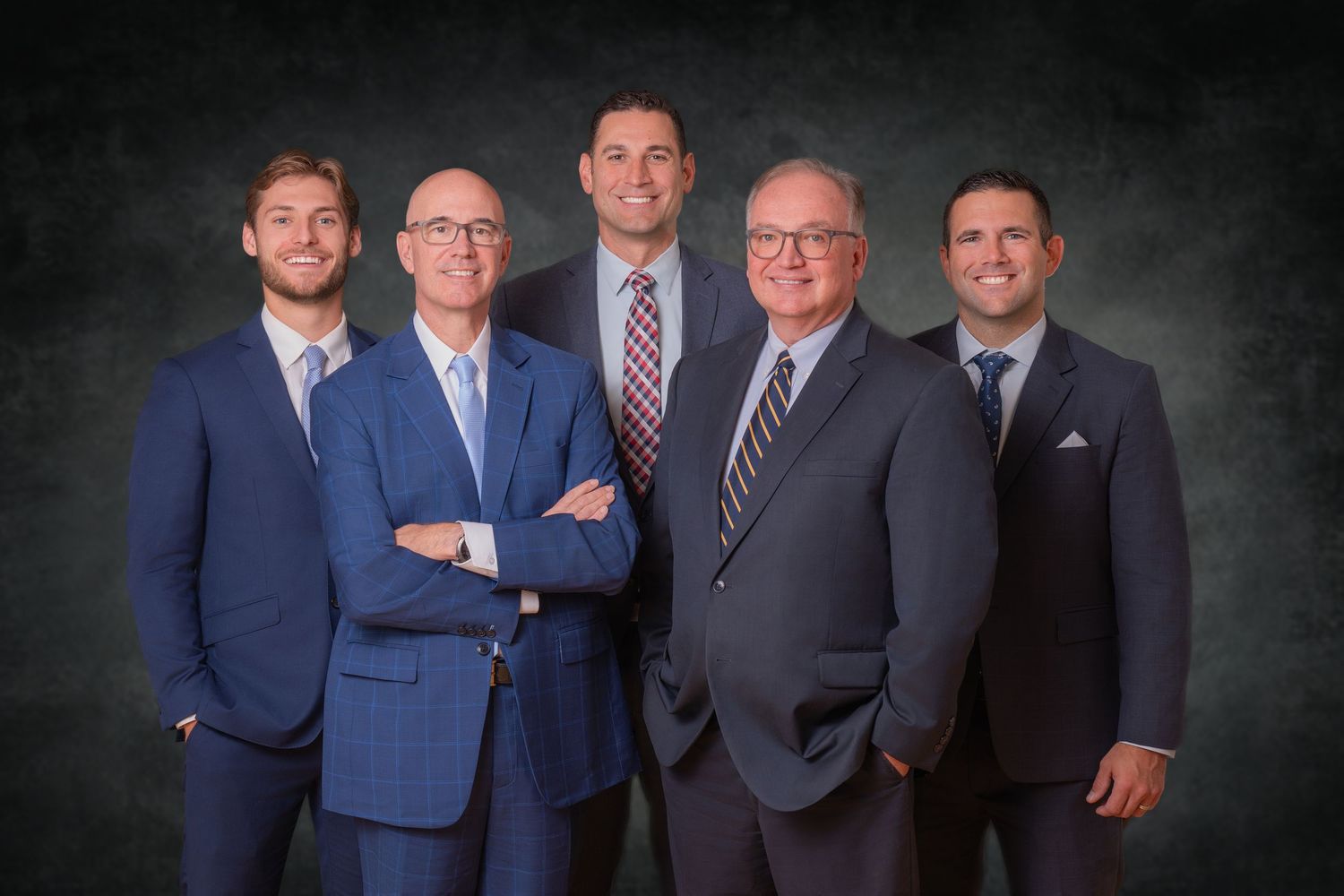 Five professional men in blue suits posing together for a formal group portrait on dark background.