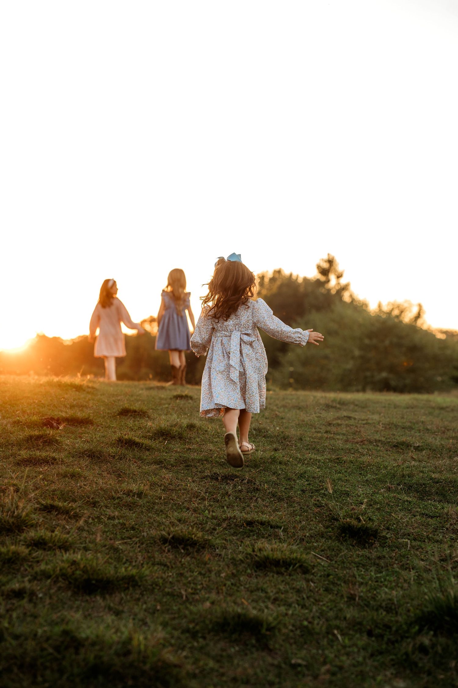 Three girls in light dresses running across a green field toward a bright sunset.