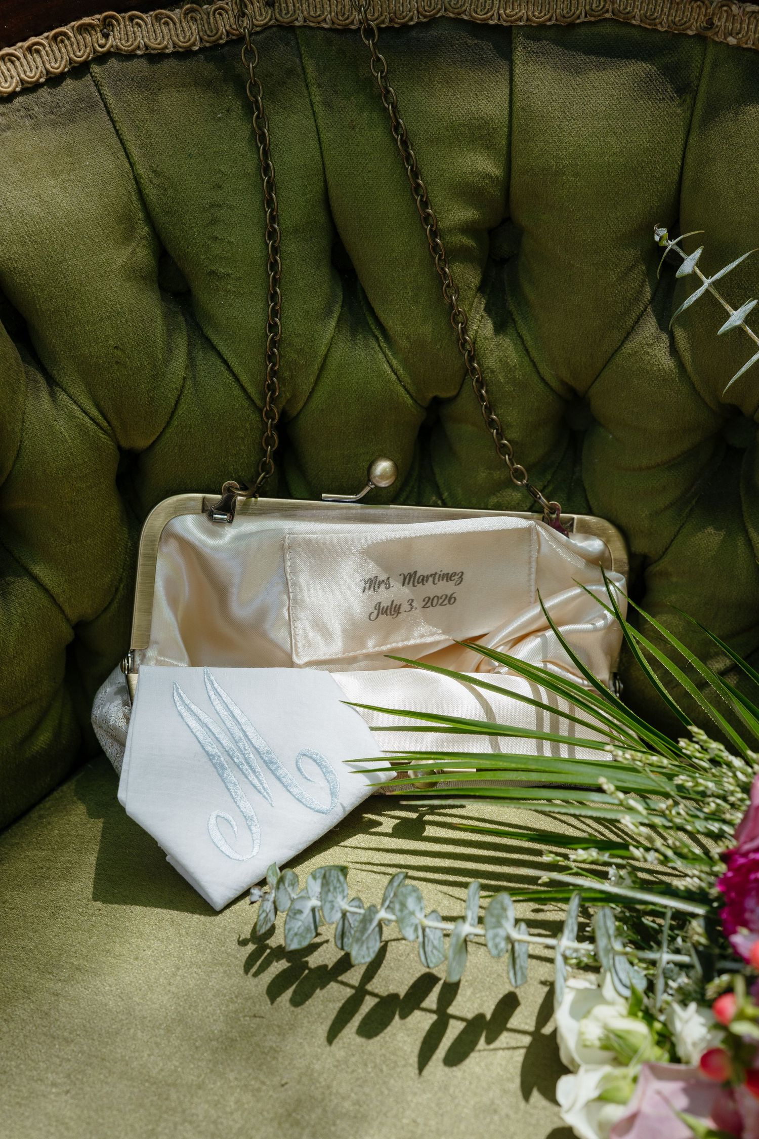 Personalized bridal clutch and monogrammed handkerchief on a green tufted velvet chair with flowers.
