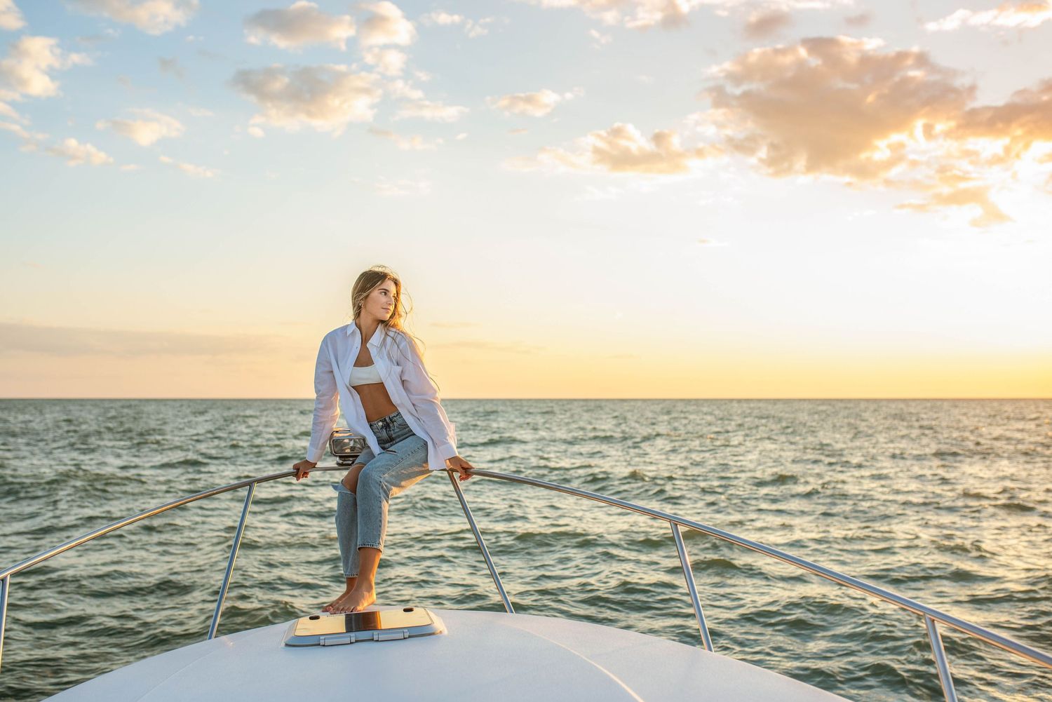 Blonde woman in white shirt and jeans standing on yacht bow at golden sunset over the ocean.