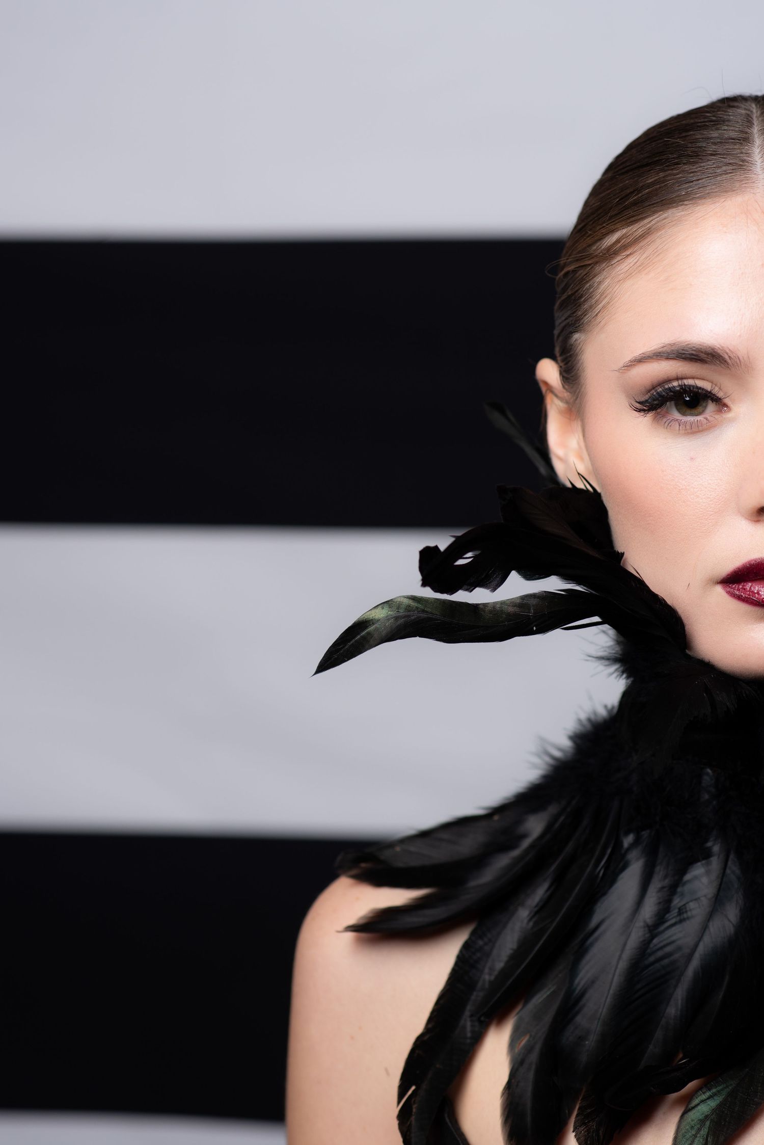 Elegant woman with dark lipstick wearing black feather accessory against striped background.
