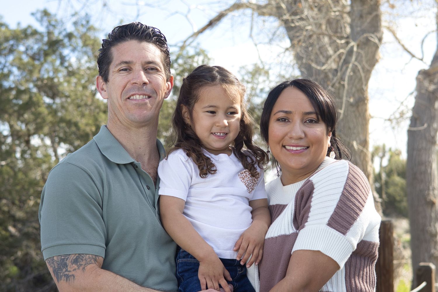 Smiling family of three poses outdoors among trees, parents holding their young daughter between them.