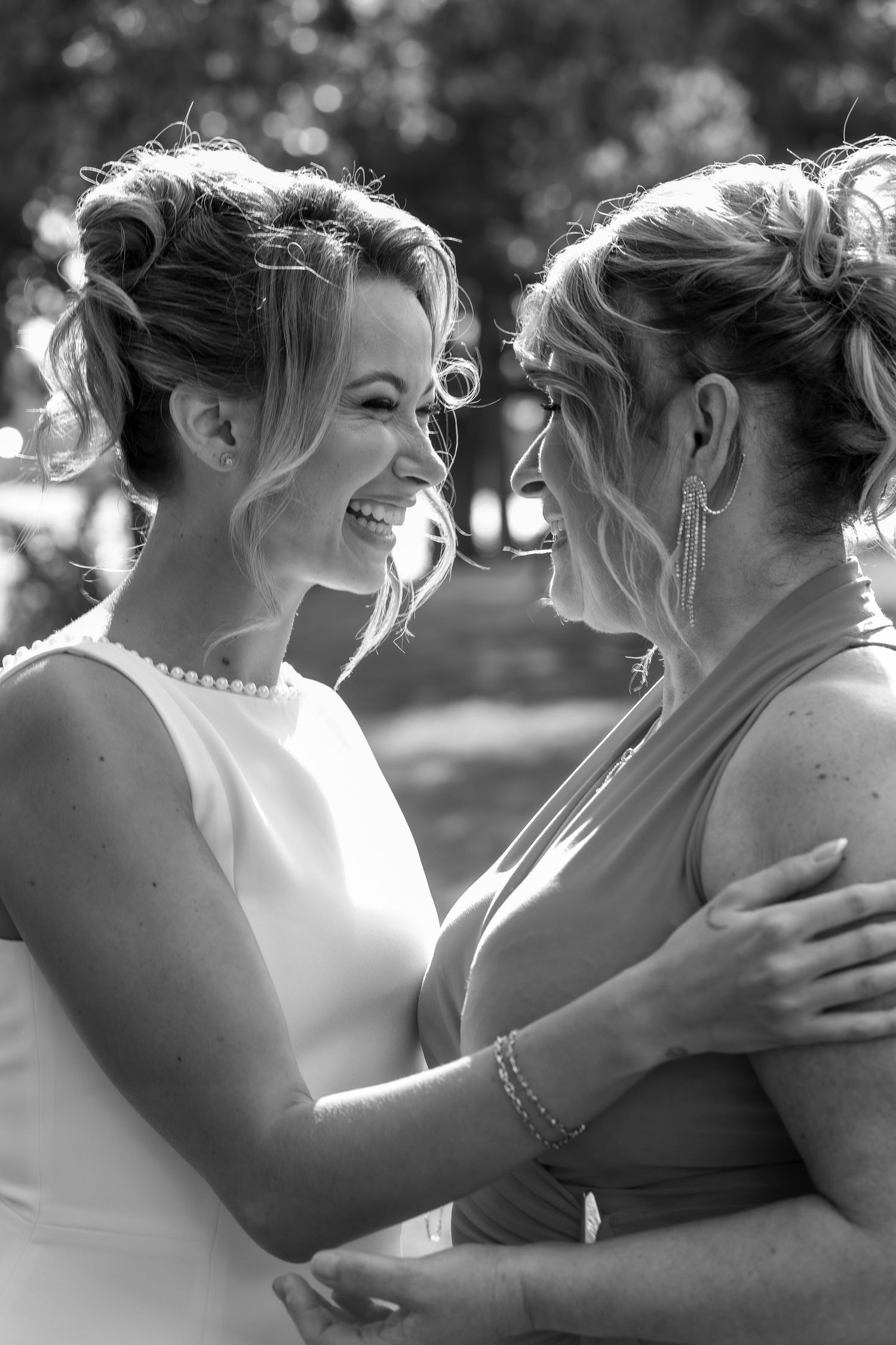Black and white photo of two smiling women in elegant updo hairstyles embracing at a wedding celebration.