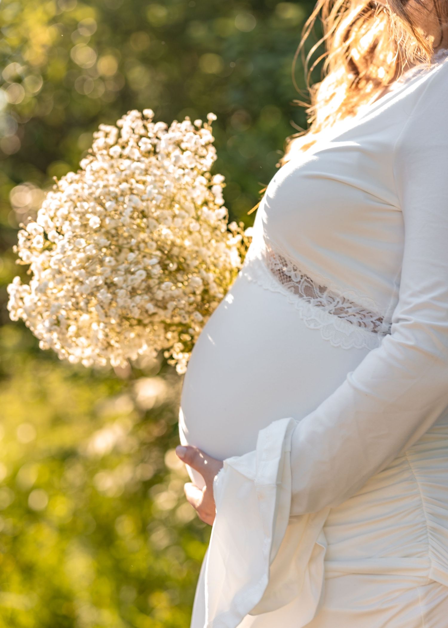 Pregnant woman in white lace dress holding baby's breath bouquet outdoors in soft sunlight.