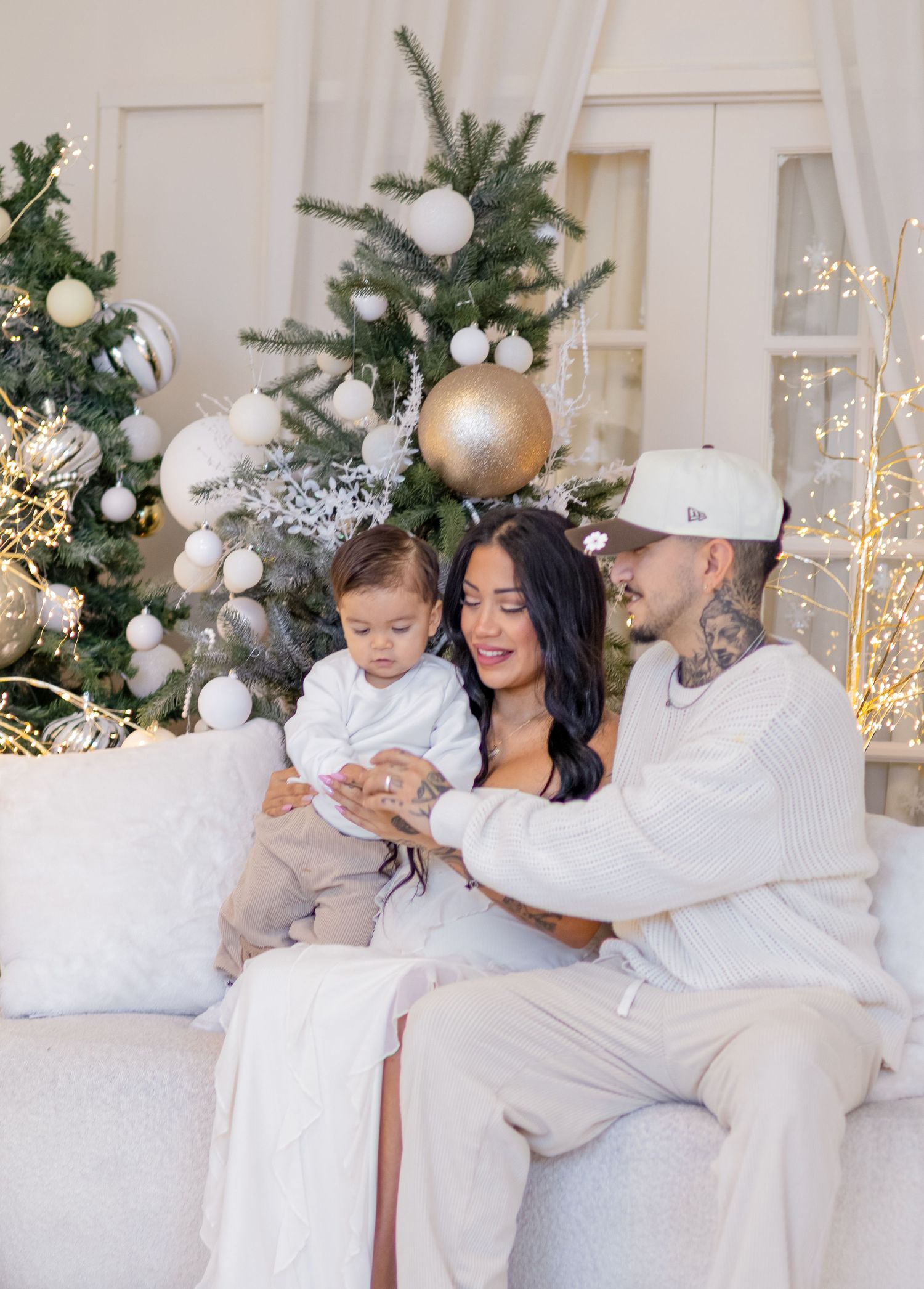 Elegant holiday photoshoot featuring mother and baby in white outfits against a Christmas-themed backdrop with ornaments.