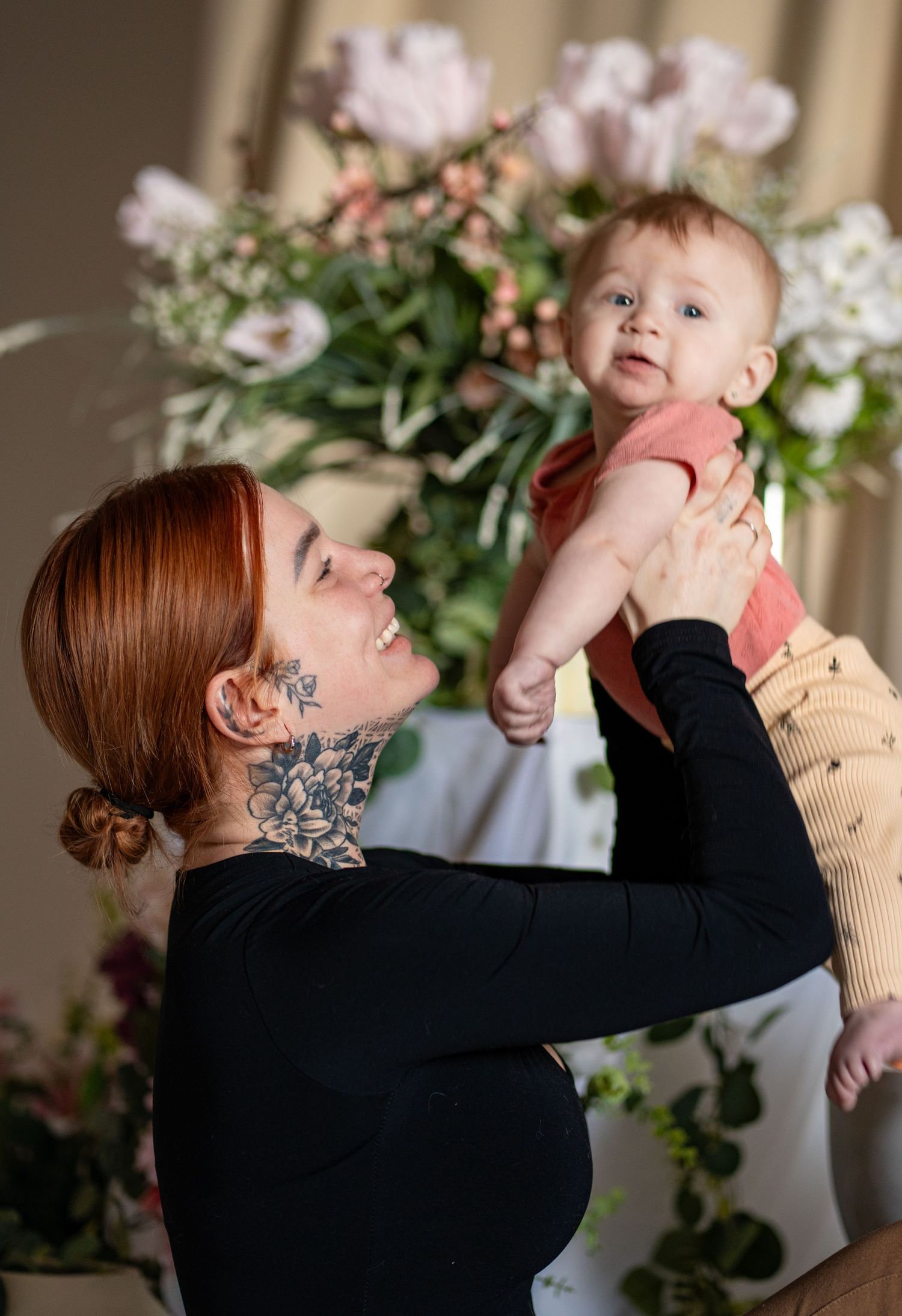 Tattooed redhead mother lifting smiling baby in red onesie with pink floral arrangement in background.