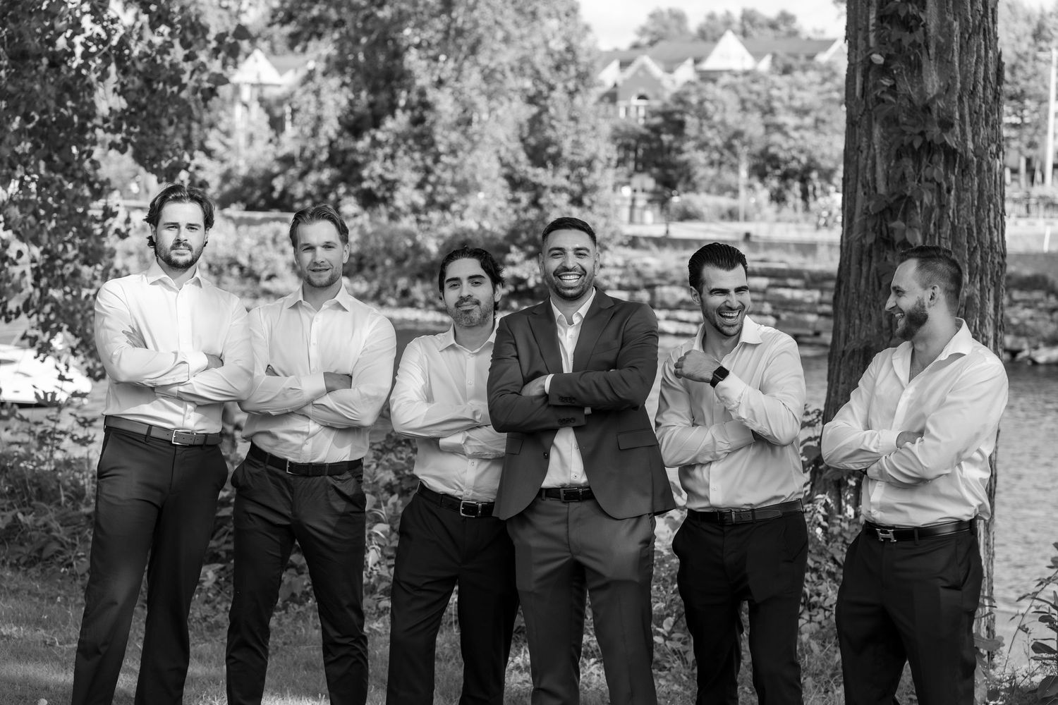 Black and white photo of six men in dress shirts smiling and posing outdoors near trees at a formal event.