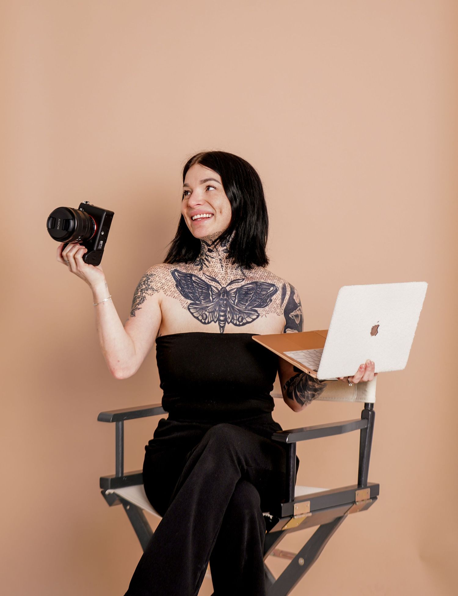 Tattooed woman in black outfit sits in director chair holding camera and Apple laptop against beige background.