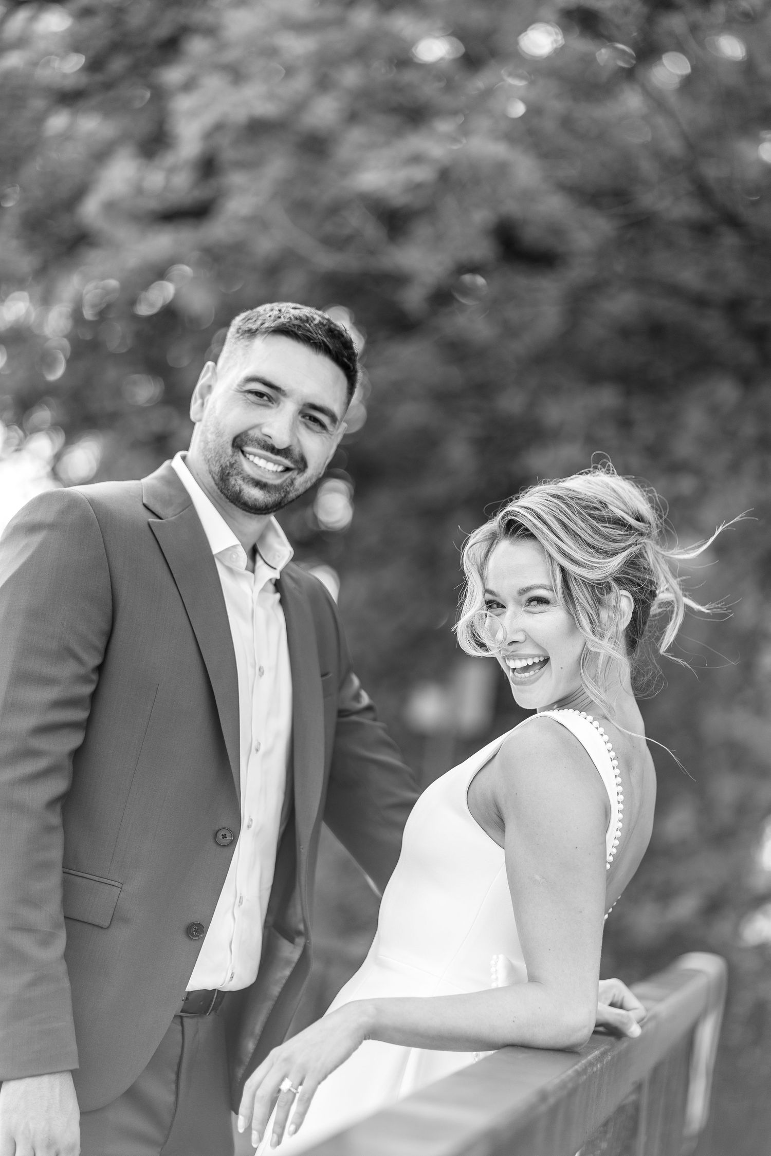 Black and white photo of a smiling bride and groom posing outdoors near a railing with lush trees in the background.