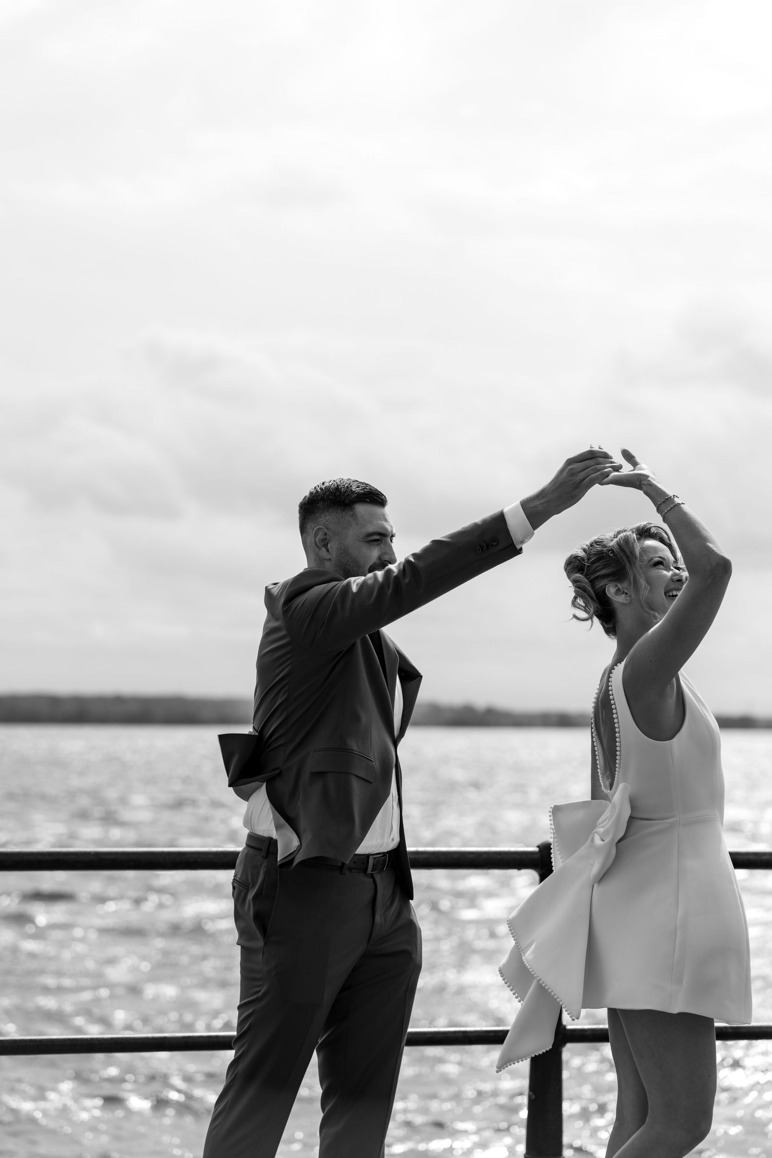 Black and white photo of a couple dancing by the waterfront, man in suit spinning woman in white dress.