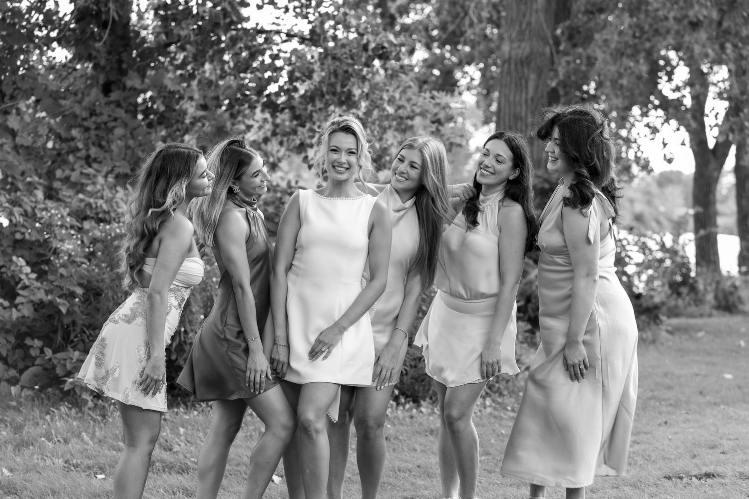 Black and white photo of a bride and six bridesmaids laughing together outdoors near trees.