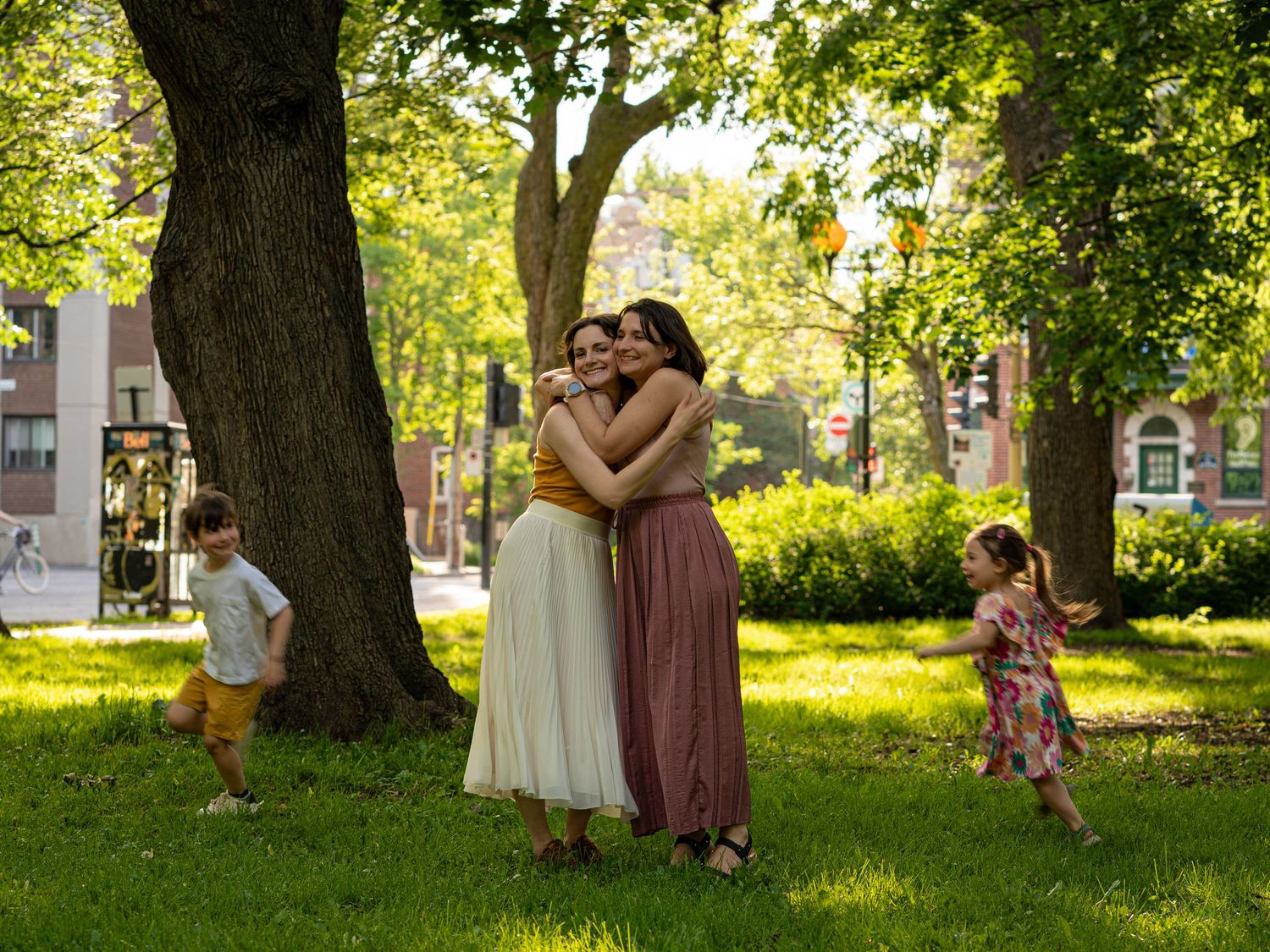 Two women hugging in a sunny park with children playing nearby under large green trees.