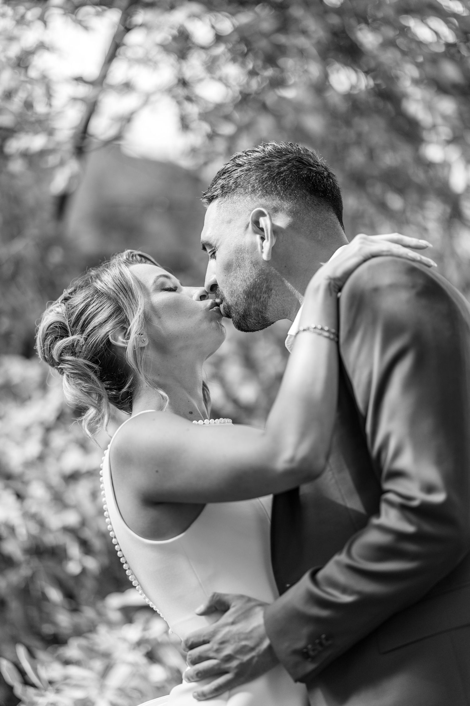 Black and white photo of a bride and groom sharing a romantic kiss outdoors, surrounded by lush greenery.