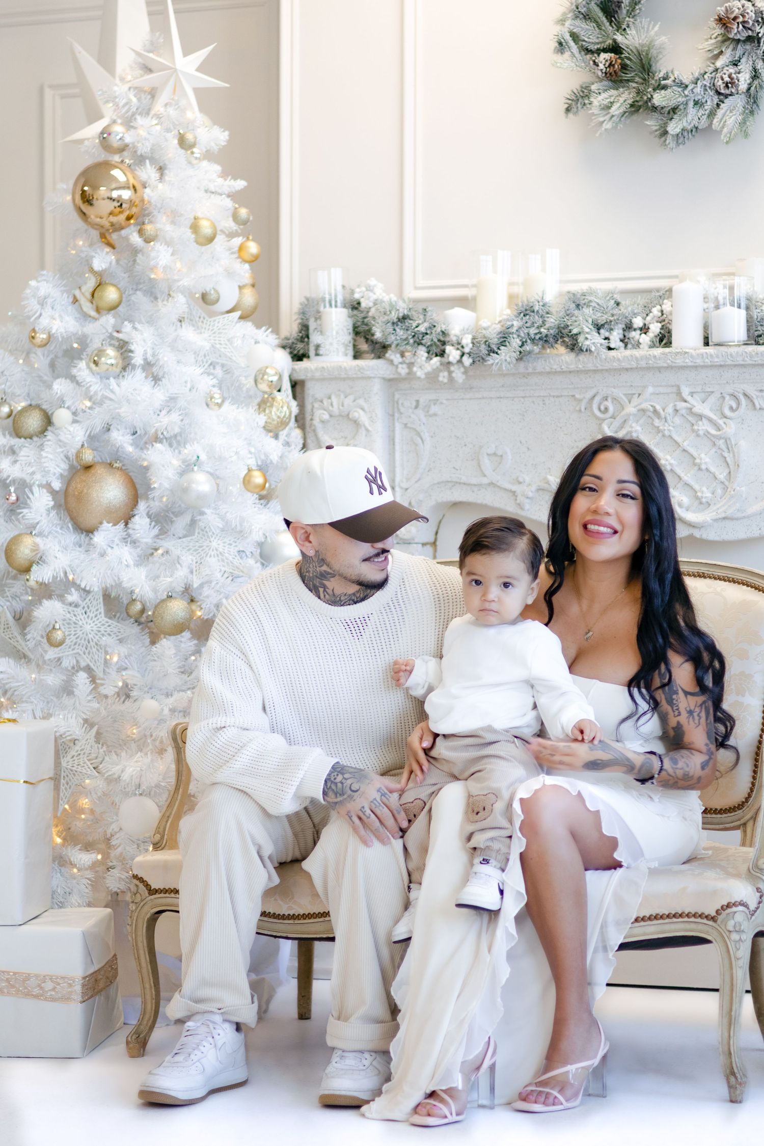 Family shares joyful moment on vintage chair beside white Christmas tree with gold decorations and wreath.