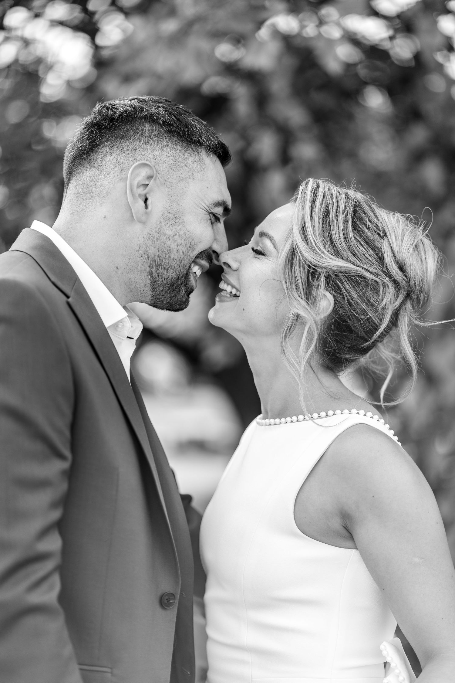 Black and white photo of a smiling bride and groom leaning in for a kiss outdoors on their wedding day.