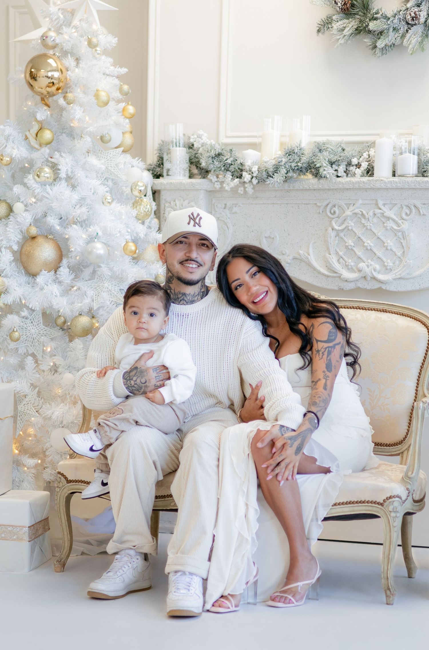 Family poses together in white outfits next to elegant white Christmas tree and gold ornaments in bright studio setting.