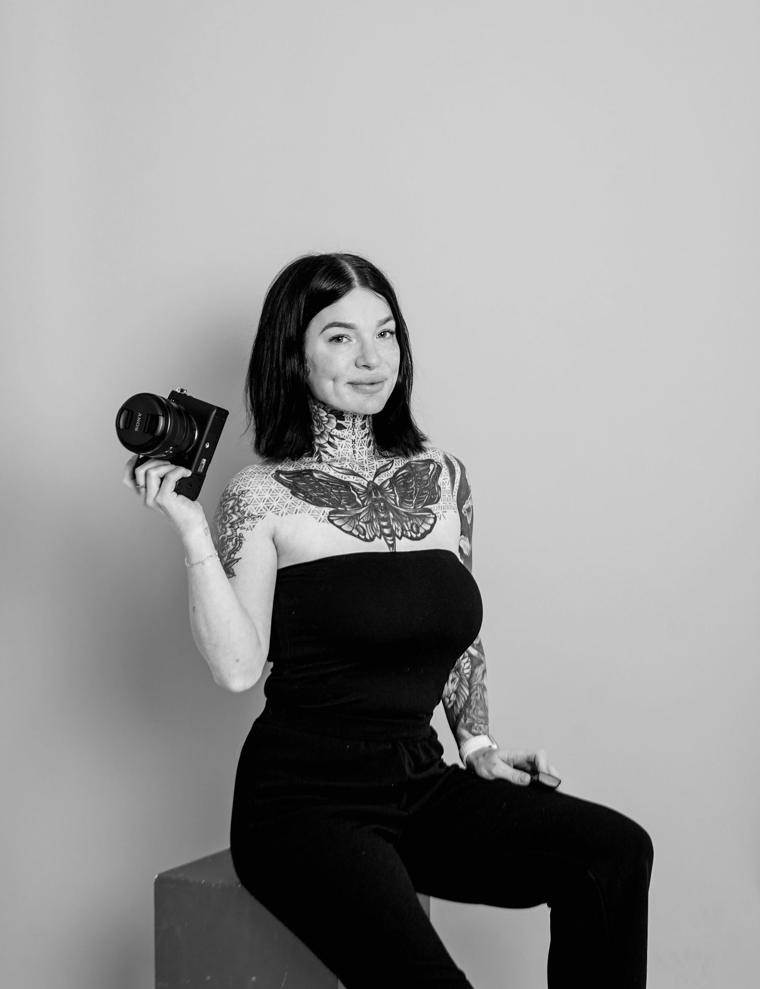 Black and white photo of a tattooed woman holding a camera, sitting confidently in a studio setting.