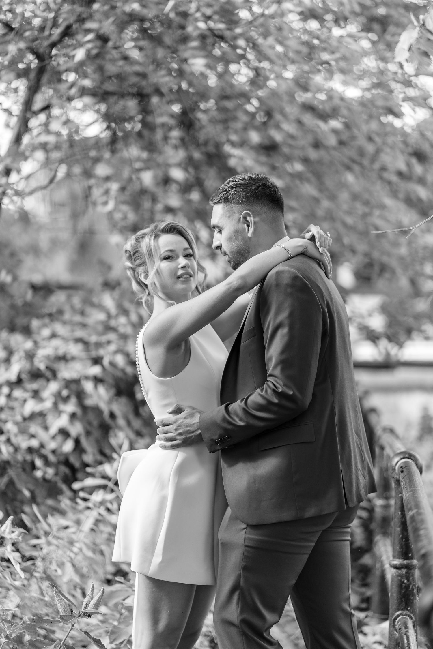 Black and white photo of a couple embracing outdoors, woman in white dress with arms around man in suit among lush foliage.