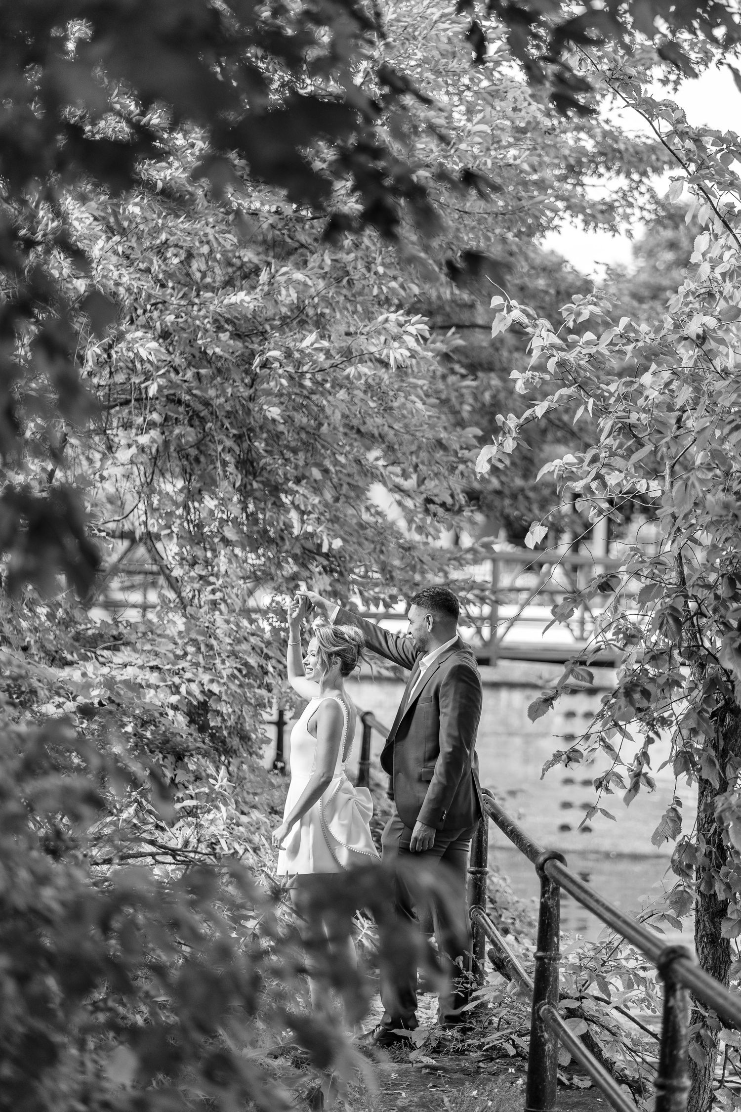 Black and white photo of a bride and groom standing among lush trees, sharing a romantic moment outdoors.