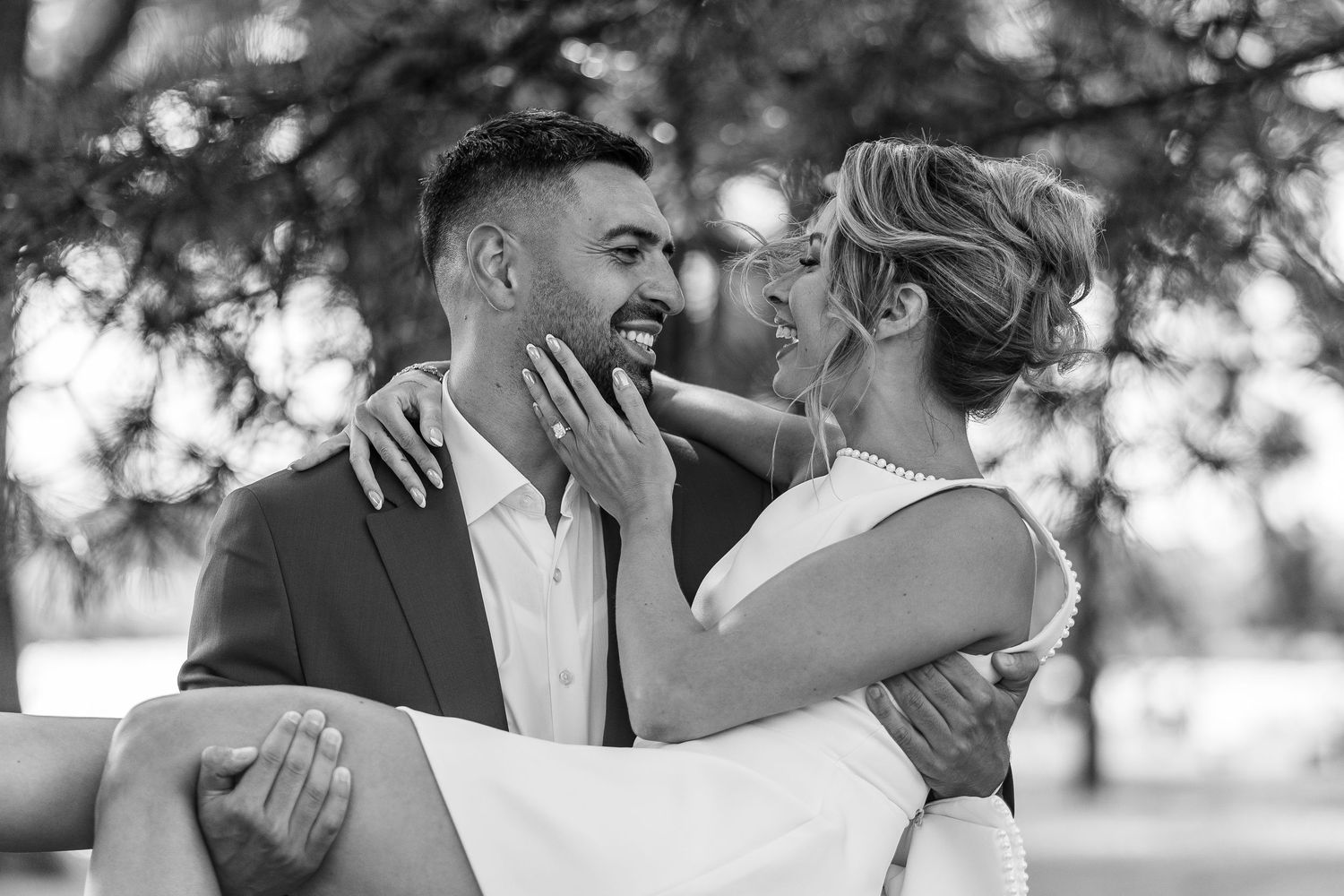 Black and white photo of a smiling couple in wedding attire embracing outdoors under trees.