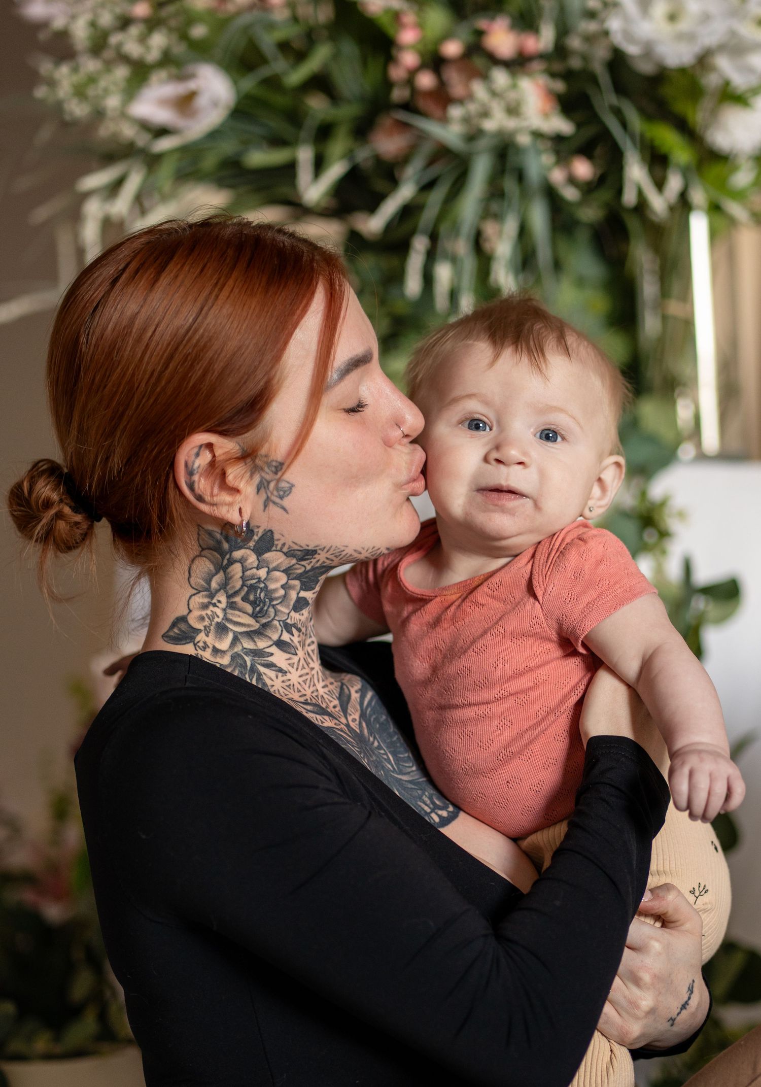 Tattooed redhead mother kissing her smiling baby in a red onesie, with floral decor in the background.