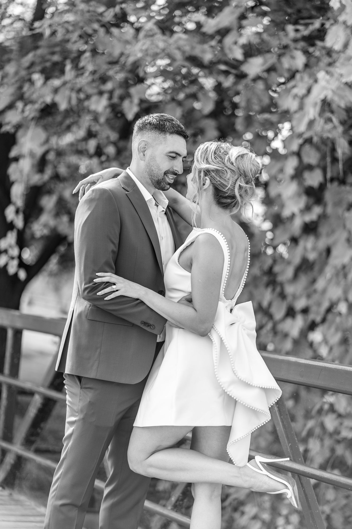 Black and white photo of a couple in wedding attire embracing on a bridge surrounded by lush greenery.