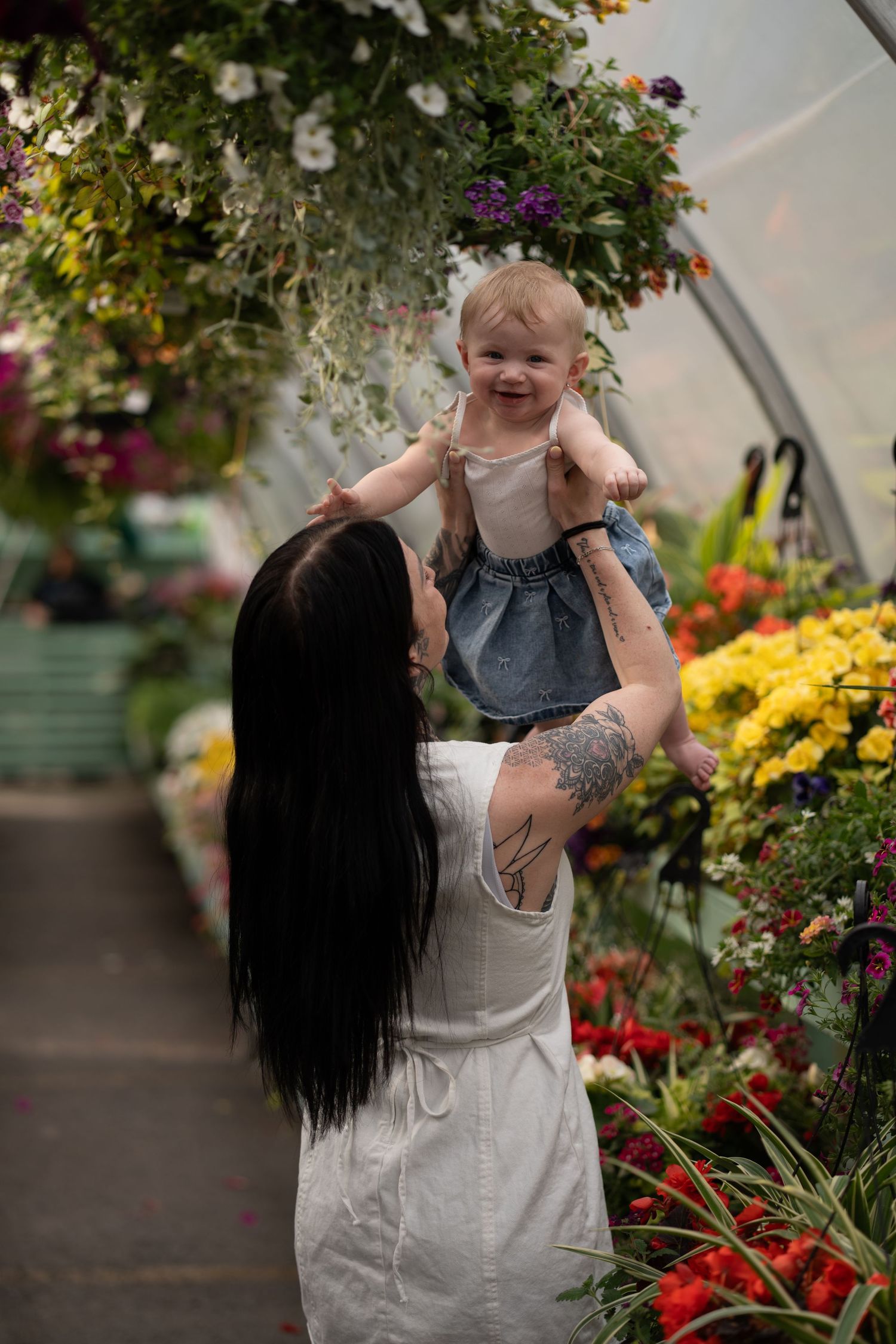 A woman with dark hair lifts a smiling baby girl amid colorful flowers in a greenhouse.