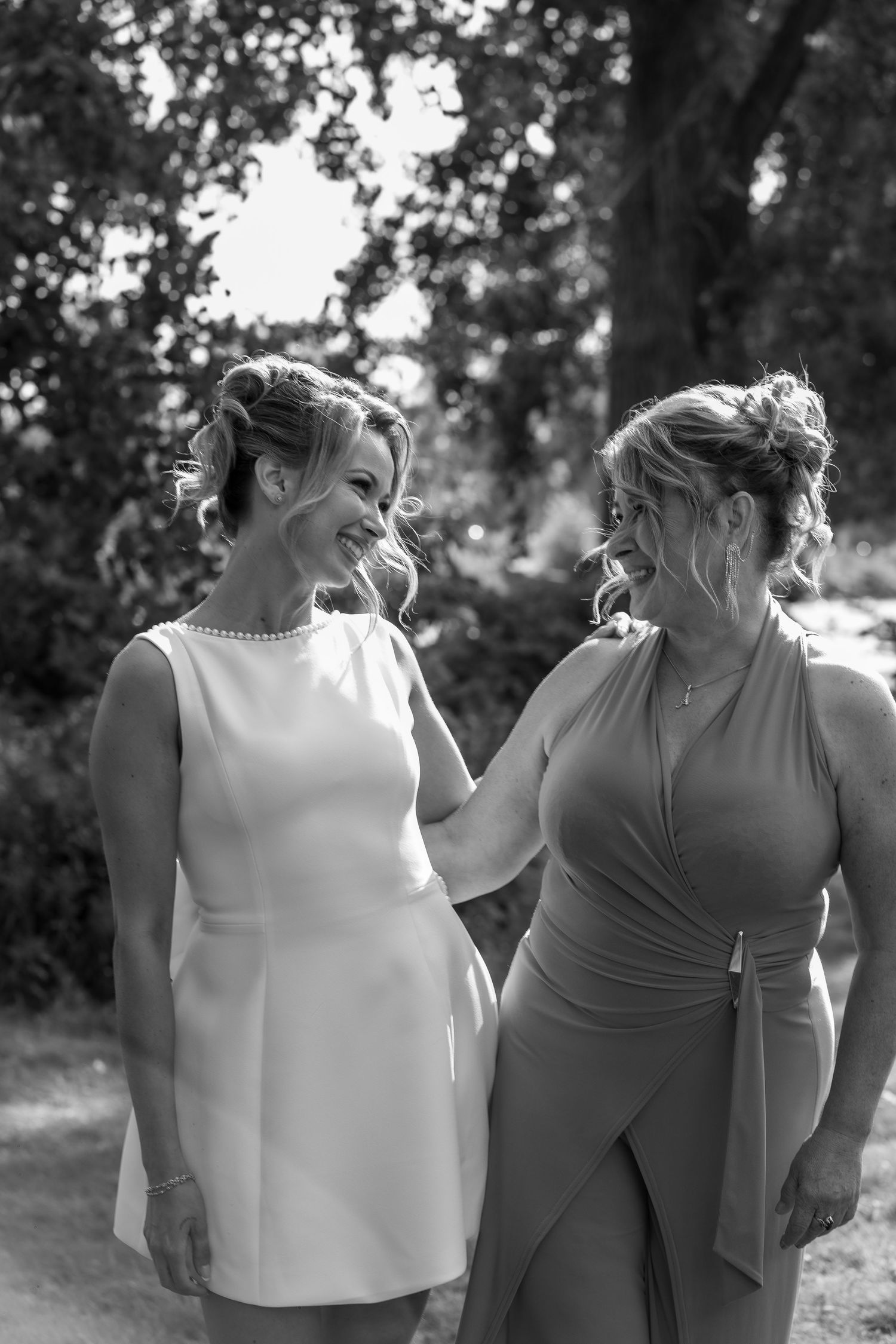 Black and white photo of two smiling women in elegant dresses sharing a joyful moment outdoors at a wedding celebration.