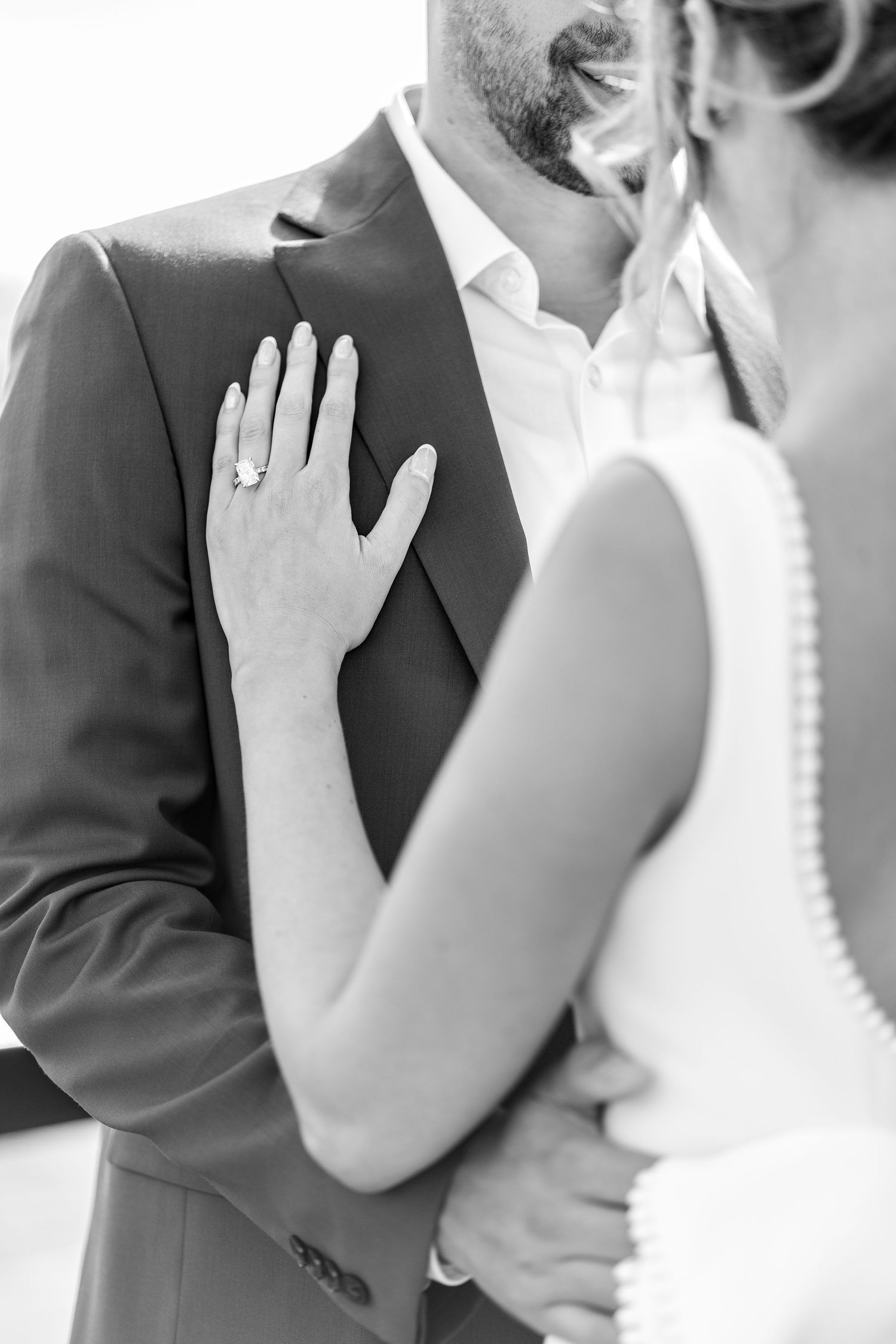 Black and white photo of a bride and groom embracing, her diamond ring visible on his chest.