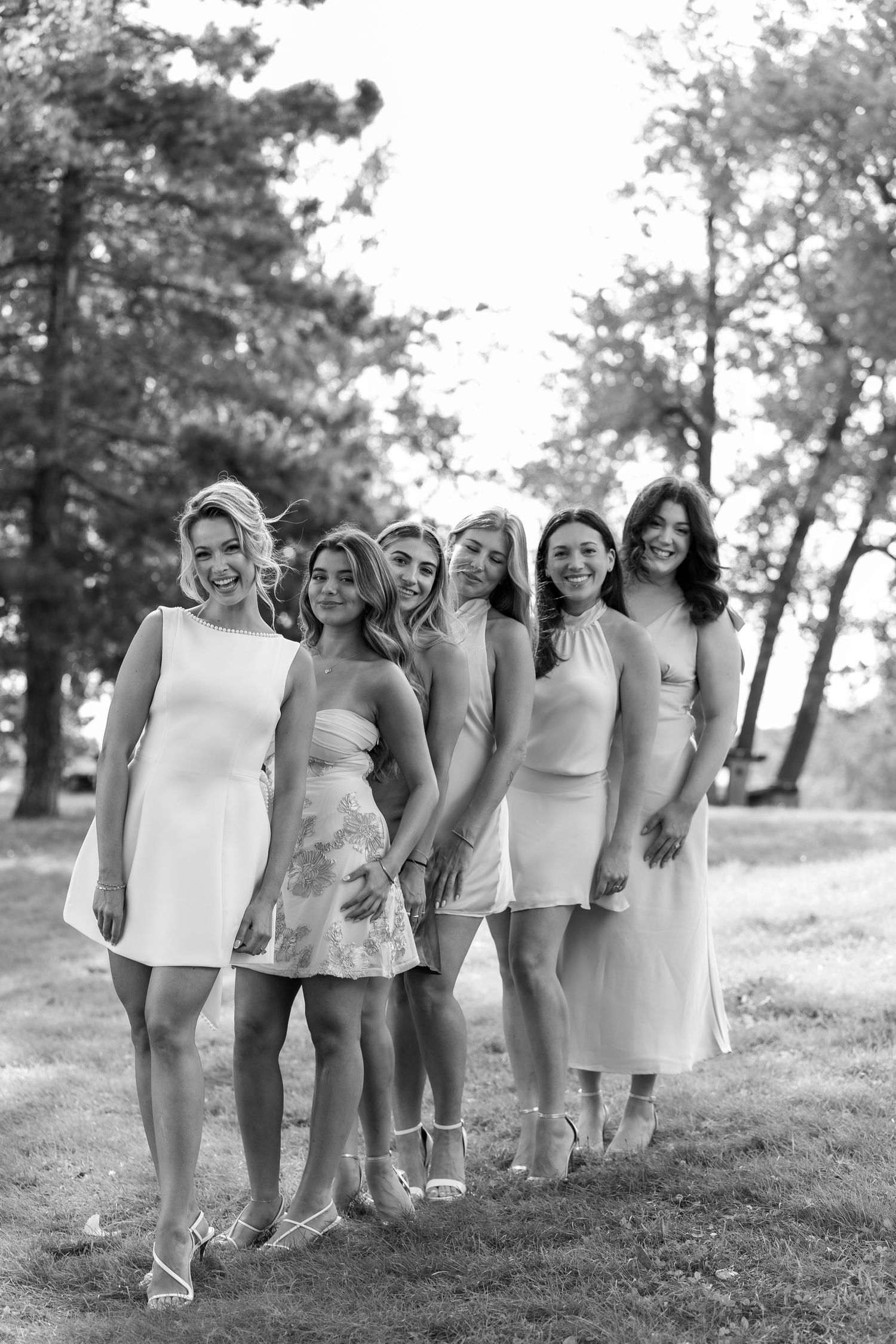 Black and white photo of six women in elegant dresses posing together outdoors in a park setting.