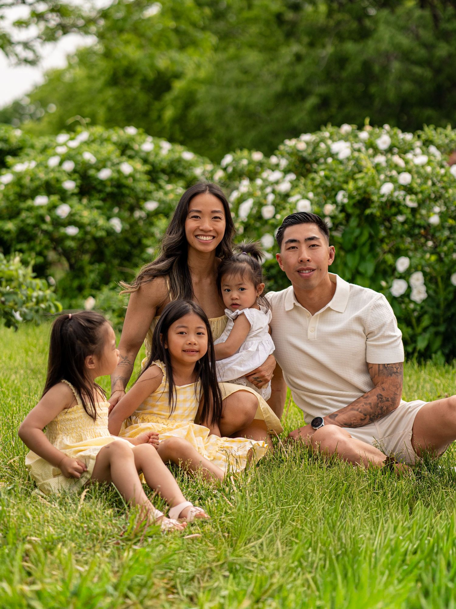 A happy Asian family of five sitting on green grass with white flowering bushes, smiling outdoors in a lush garden setting.