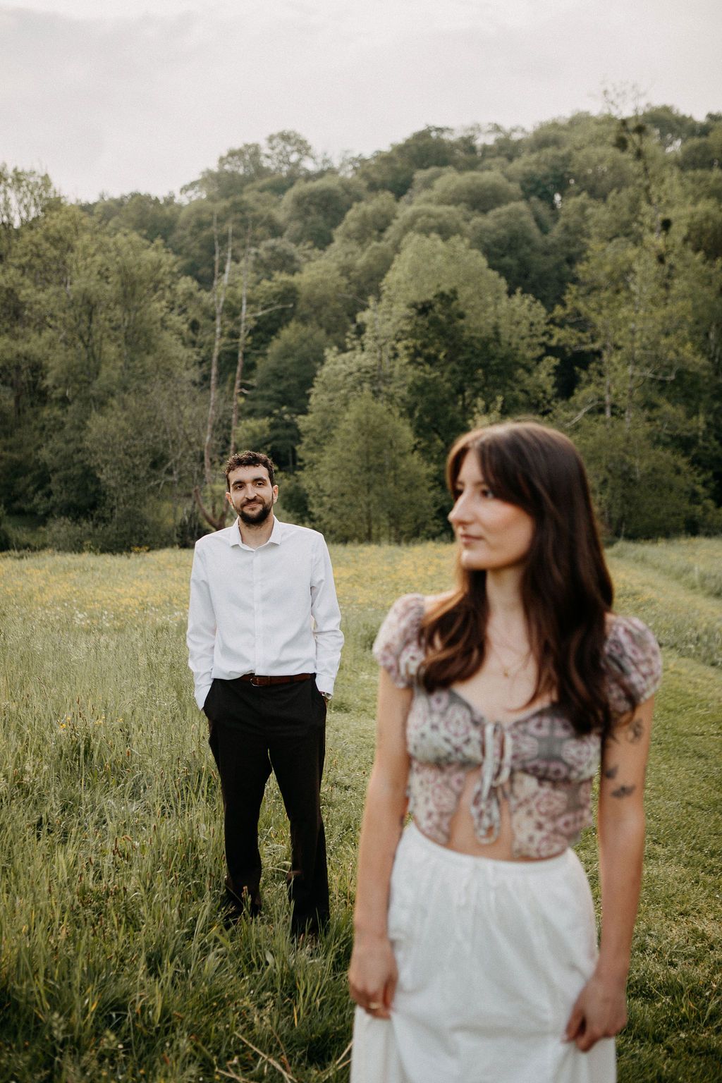 A man in a white shirt gazes at a woman standing in a lush green meadow landscape.