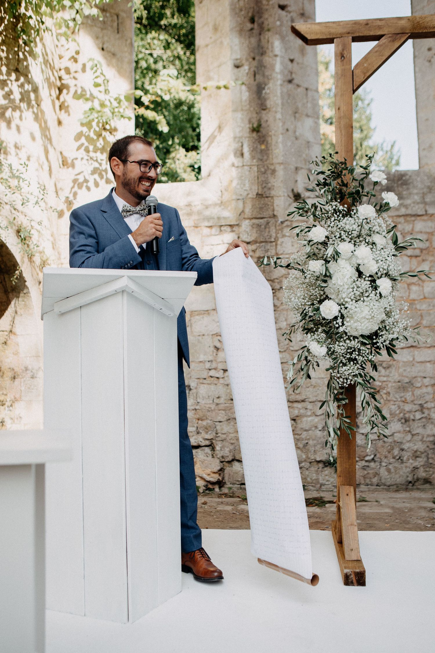 Officiant speaking at a white podium during an outdoor wedding ceremony in a rustic stone venue with floral arch.