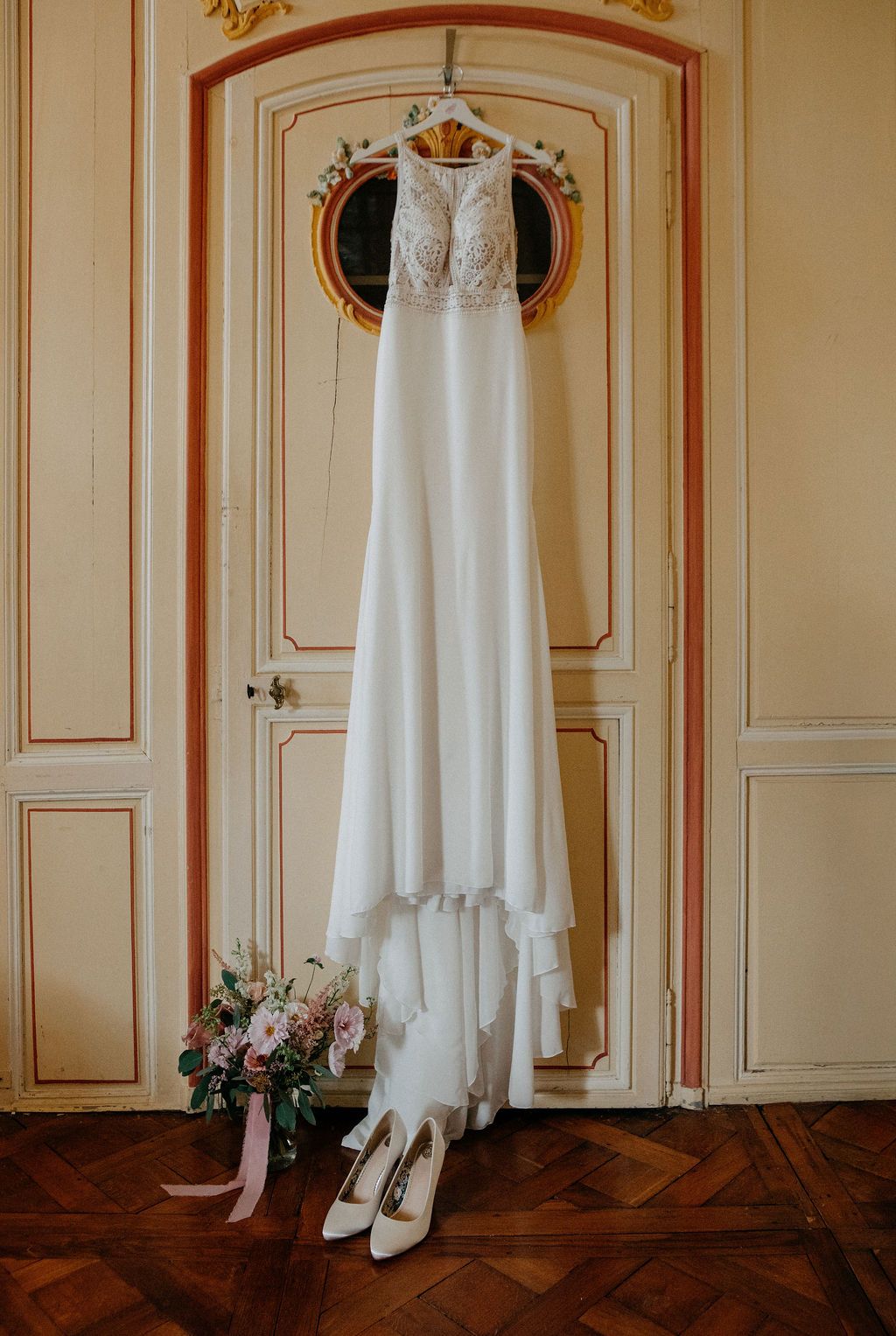 Elegant white lace wedding dress hanging on a door with bridal bouquet and heels below.