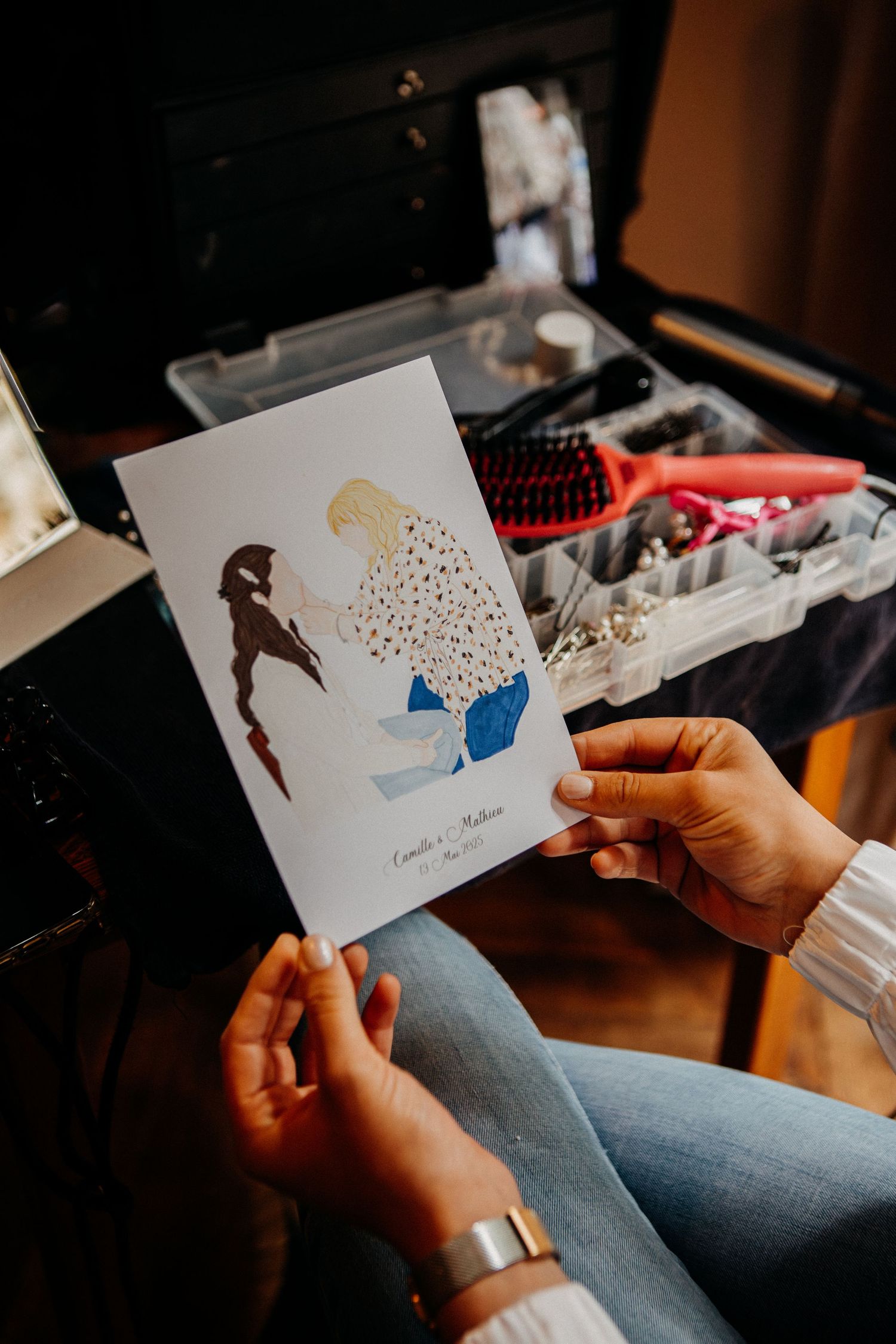 Bride holding a custom illustrated wedding card showing two figures, with a makeup vanity and hair tools in the background.